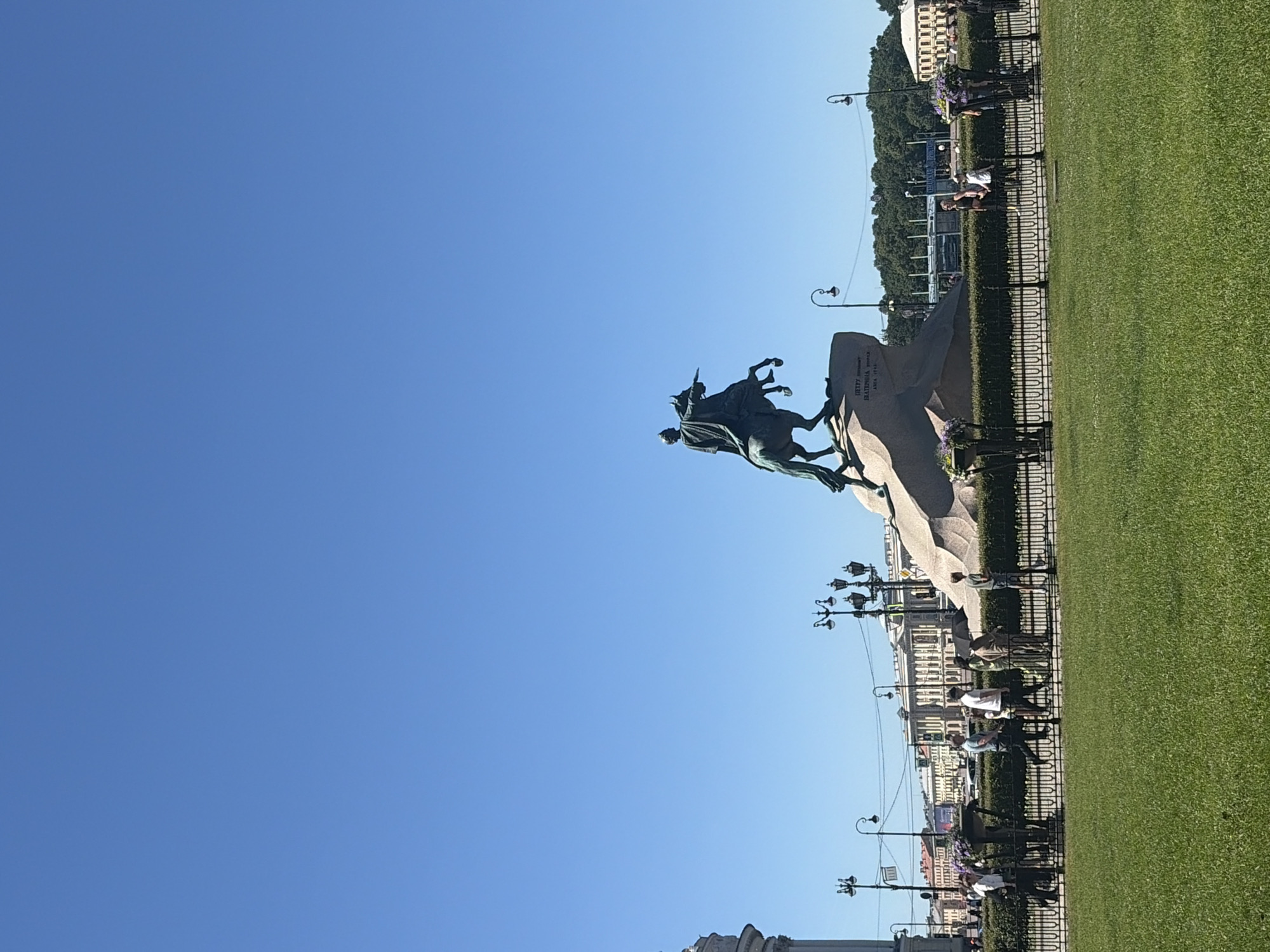 This image captures a daytime view of the Bronze Horseman, a monument to Peter the Great, located in Senate Square in Saint Petersburg, Russia. The monument, a dark bronze equestrian statue, is positioned on a large rock pedestal under a clear, bright blue sky. In the foreground, a meticulously manicured green lawn stretches across the scene. A black metal fence separates the lawn from a paved walkway where several individuals are walking. The people are casually dressed, suggesting a pleasant, likely warm day. Behind the fence and walkway, a dense green hedge runs horizontally, with decorative planters filled with flowers at intervals. In the background, classic Saint Petersburg architecture with light-colored facades and numerous windows lines the square. Streetlights with multiple lamps are visible, along with trees and a section of what appears to be a modern structure with a blue roof. The overall atmosphere is one of a public square on a sunny day, with tourists and locals enjoying the historical landmark and its surroundings.