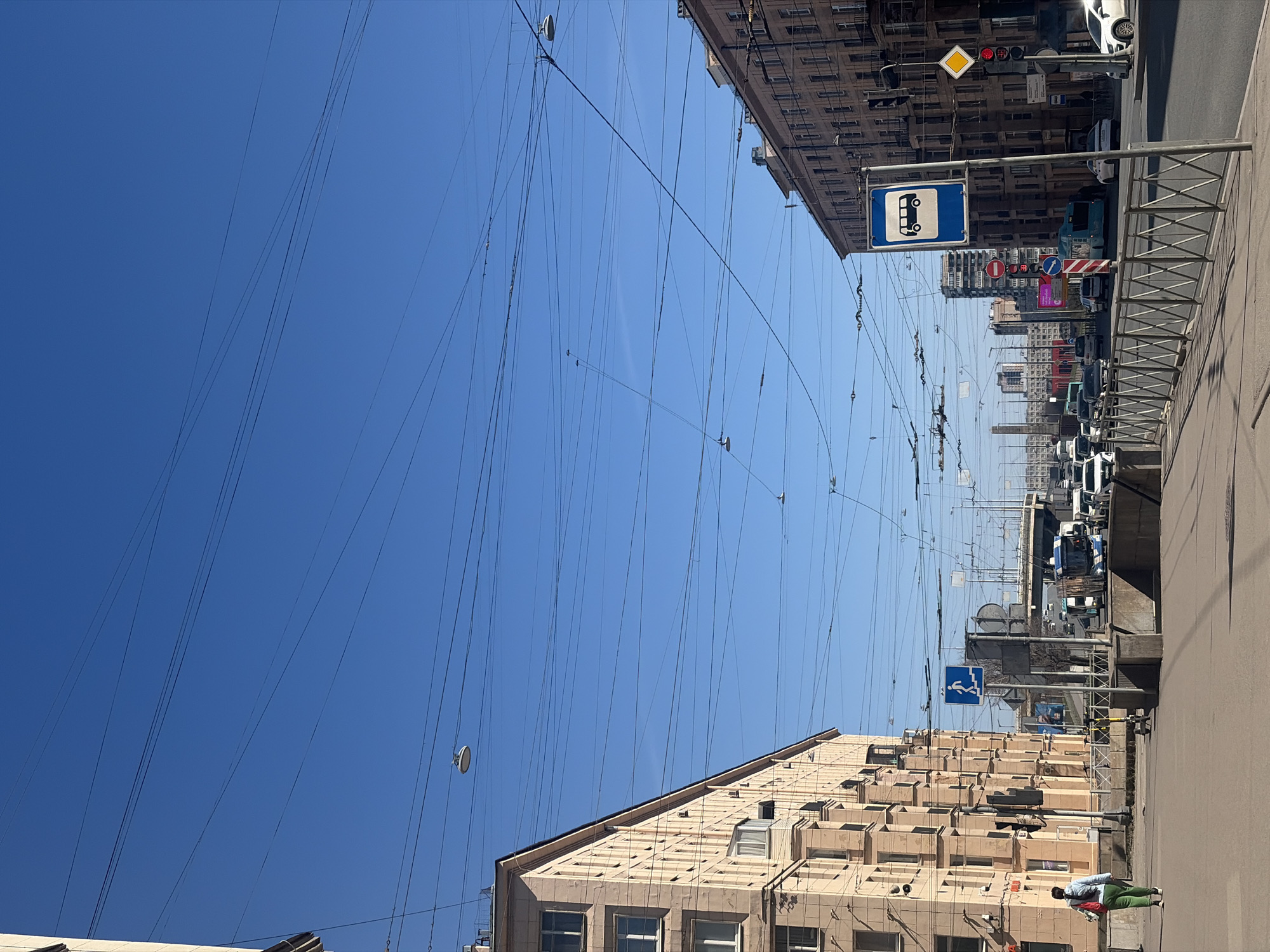 The image captures a street in Saint Petersburg, Russia, viewed from a low angle, with a clear, blue sky visible above. Power lines crisscross overhead, creating a web-like pattern against the sky. Buildings line both sides of the street, constructed of light-colored stone or concrete, with rectangular windows and some projecting architectural details. A bus stop sign is visible on the right. Traffic lights and various street signs are also visible, along with metal barriers lining the pedestrian walkways. Several vehicles are parked along the street, and a pedestrian is walking on the sidewalk. The street appears to be paved and generally clean.