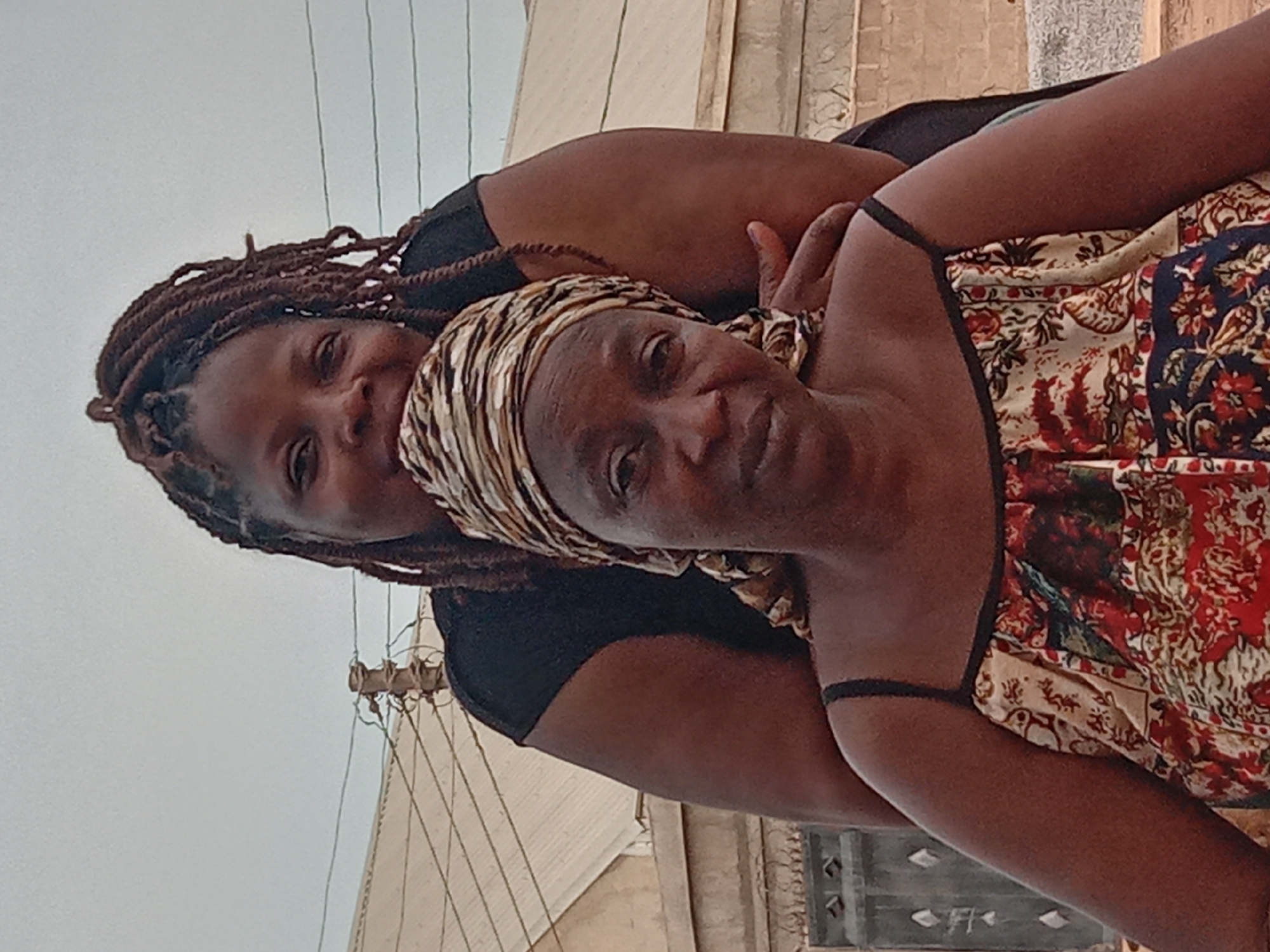 Two women are embracing. The younger woman has brown braids and is wearing a black top. The older woman is wearing a patterned dress and a patterned headscarf. The location appears to be in Sabon Tasha, Nigeria, given the visible architecture and environment. Wires are visible in the sky.