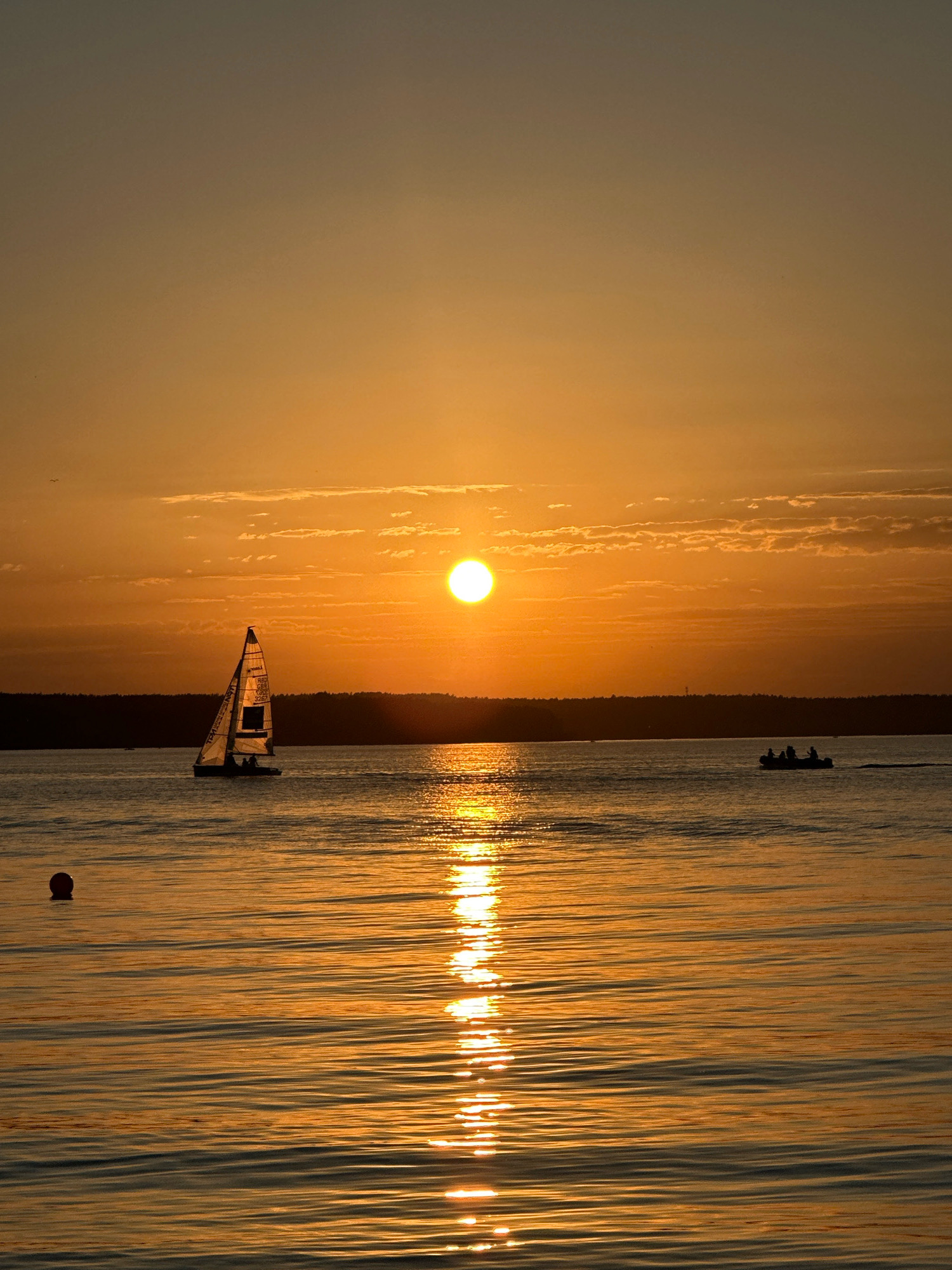 This is a vertical, eye-level, medium shot photograph taken at sunset on a body of water. The sun, a bright orb, is positioned in the upper center of the frame, casting a warm orange glow across the sky and water. The sky transitions from a light gray at the top to a rich orange near the horizon, with some scattered, wispy clouds.
In the middle ground, a sailboat with its sail raised is visible on the left, silhouetted against the bright sunset. The boat appears to have at least two people on board. Further to the right, a small motorboat with three occupants is also in motion. A dark, treed shoreline is visible in the distance, forming a distinct horizon line.
The foreground is dominated by the water, which reflects the vibrant colors of the sunset, creating a shimmering path of light extending from the horizon towards the viewer. On the left side of the frame, a small, dark red buoy is floating on the water. The water's surface is slightly rippled, indicating a gentle breeze.
The location is identified as a lake or large river within the Ждановіцкі сельскі Савет, Belarus, likely during the summer months given the warm lighting and recreational activities depicted. The overall mood of the image is serene and tranquil, capturing the beauty of a sunset over water.
