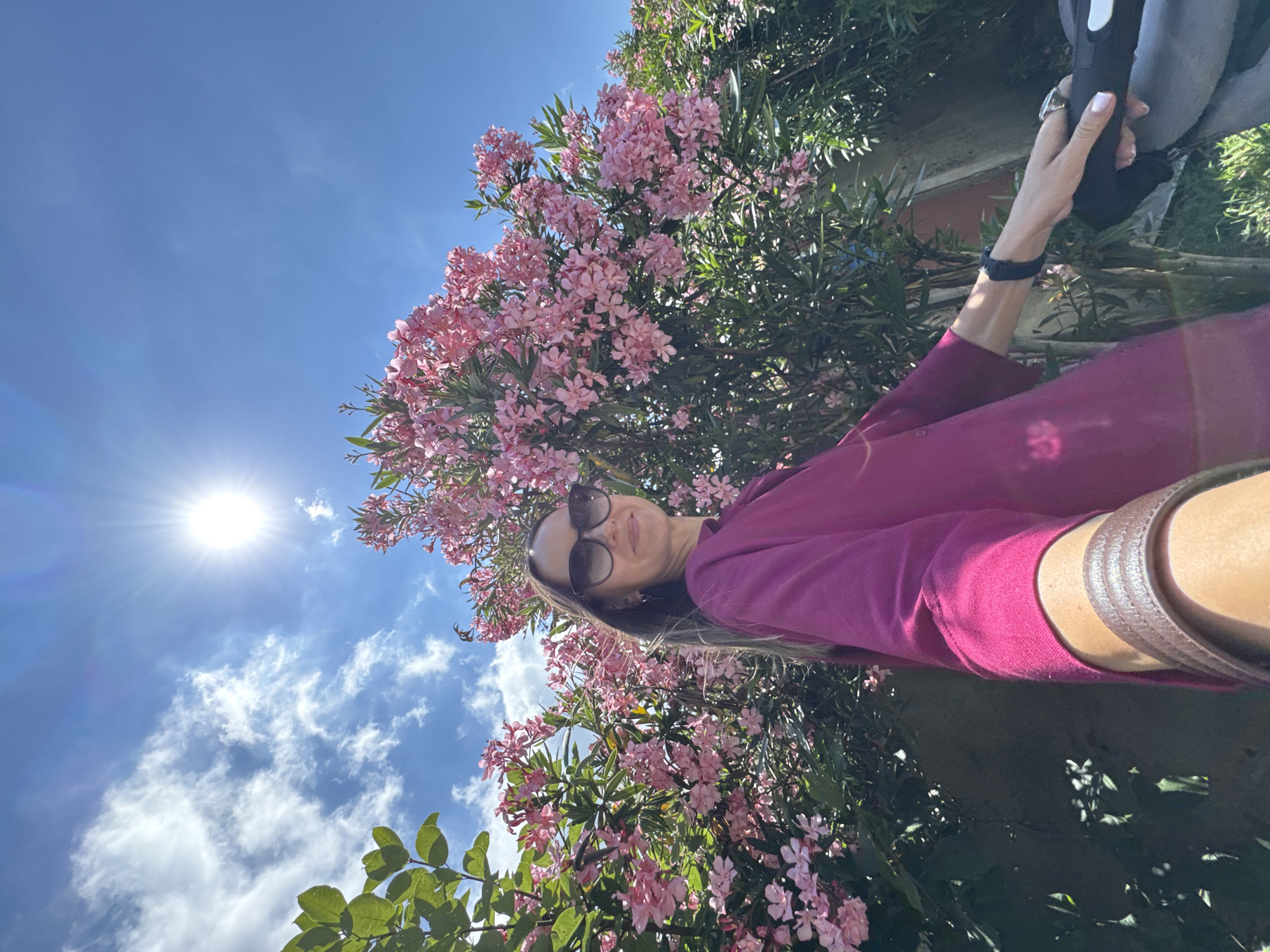 The image shows a person taking a selfie outdoors in front of a blooming tree with pink flowers. The person is wearing sunglasses and a magenta top, and has a brown strap going across their shoulder. The background consists of a bright blue sky with some white clouds and the sun, which is shining brightly. Green leaves from the tree are also visible. The scene suggests it is a sunny day, possibly in the late morning or early afternoon, given the angle of the sun. The blooming flowers and green foliage hint at a warm weather season, like spring or summer. The setting appears to be a garden or park. There are no visible text elements in the image. The orientation of the photo suggests it was taken in portrait mode and then rotated.