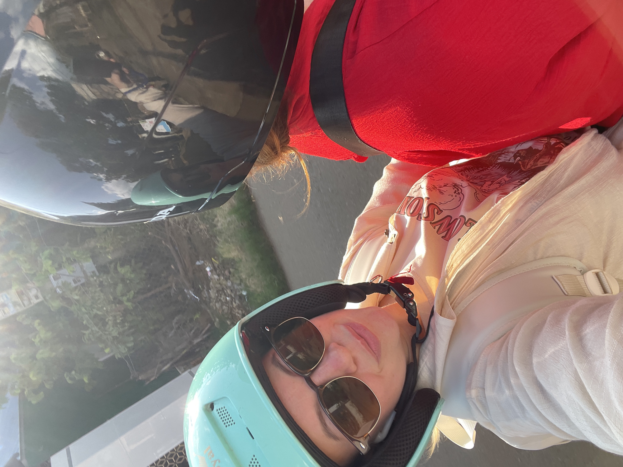 Two individuals are depicted riding a motorbike in Thôn 4, Vietnam. The person in the foreground, facing the camera, is a female wearing a light teal open-face helmet with "Royal" text visible and dark-lensed sunglasses with a brown frame. Her mouth is closed with a slight upward curve. She is wearing a light-colored, long-sleeved outer garment over a t-shirt displaying a graphic design with partially visible text "WSON" and imagery of mountains and animals. A light-colored backpack is worn over her outer garment. The person seated in front of her, facing away from the camera, is wearing a glossy black full-face helmet with "HONDA" visible on the side. The black helmet reflects a cloudy blue sky, trees, and a partial reflection of the individual holding the camera. This person is wearing a bright red long-sleeved garment, with a dark strap (possibly from a backpack or safety restraint) extending horizontally across their upper back. Light-colored hair is visible beneath the back edge of their helmet, illuminated by direct sunlight. The scene is outdoors under strong sunlight, casting highlights and some lens flare from the upper left. The background features a paved road, green foliage, trees, and a light-colored building with a patterned section on the left.
