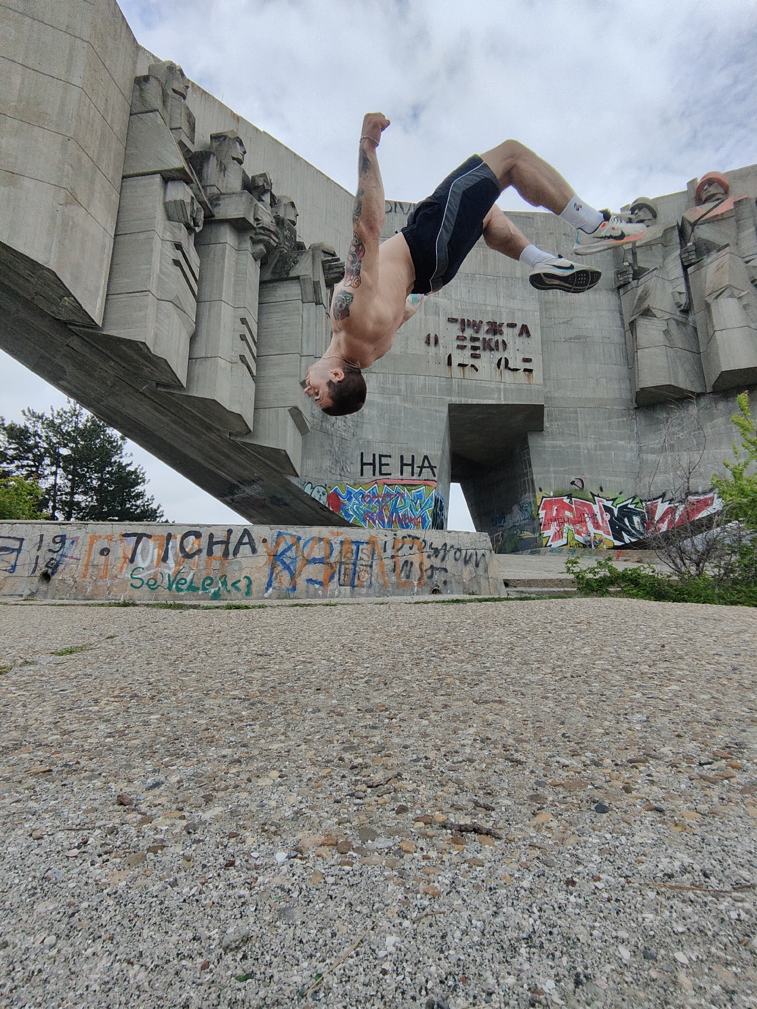 A low-angle wide shot shows a shirtless man in mid-air performing a backflip in front of the Monument to the Bulgarian-Soviet Friendship in Varna, Bulgaria. He is wearing black shorts with a white stripe, white socks, and running shoes. He is facing the camera and his left arm is raised above his head. His body is curved in an arc, with his head pointing down towards the graffiti-covered concrete barrier in the midground. Behind the barrier is the concrete monument. The ground in the foreground is covered with pebbles. The sky is overcast.