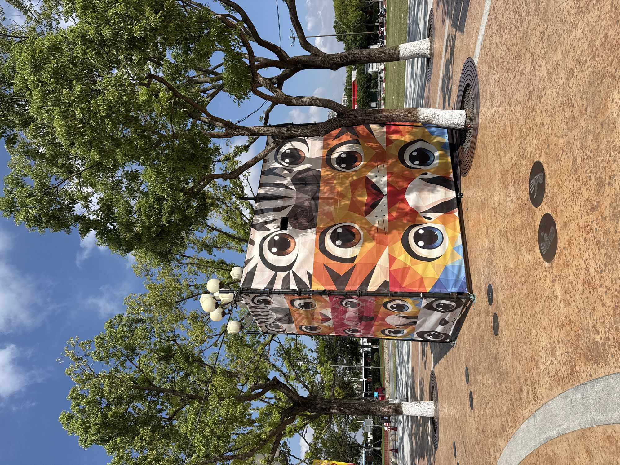 An outdoor rectangular art installation, featuring a collage of cartoon animal faces arranged vertically, is positioned in a paved public space in Pudong, China. The installation has a checkered pattern of tiger, zebra, and panda faces, each composed of large, expressive eyes and geometric shapes. The art structure is surrounded by trees with white-painted trunks and a light blue sky with scattered clouds. The ground is paved with a patterned reddish-brown material, and dark circular imprints, possibly from previous events, are visible on the pavement. In the background, a street with vehicles and pedestrians, as well as buildings and more greenery, can be seen.