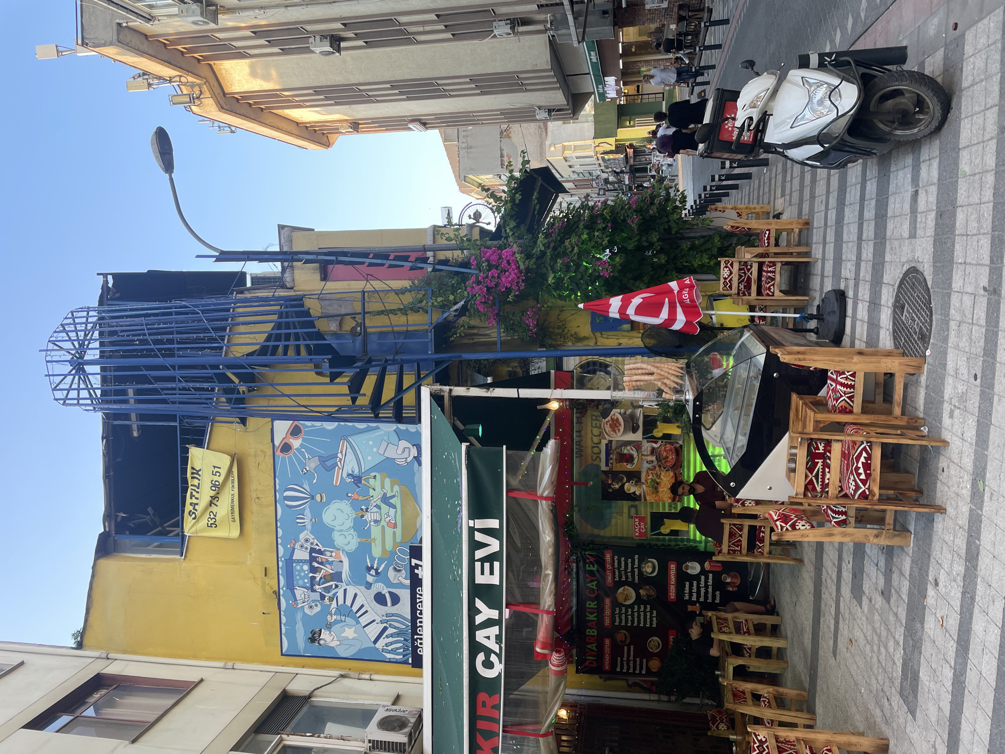 This image depicts a street scene in Kadıköy, Turkey, featuring an outdoor seating area of a cafe called "DİYARBAKIR ÇAY EVİ." The cafe has green awnings and tables with wooden chairs adorned with red and white patterned cushions. A large, colorful mural is painted on the yellow wall of the building behind the cafe, depicting a lively scene with people, hot air balloons, and musical instruments. A prominent blue spiral staircase is attached to the side of the building. In the foreground, a white scooter is parked on the sidewalk. The street is paved with grey and white tiles, and there are several streetlights and electrical wires visible. People are seated at some of the tables, and others are walking in the background. The scene is bathed in sunlight, suggesting it is daytime.