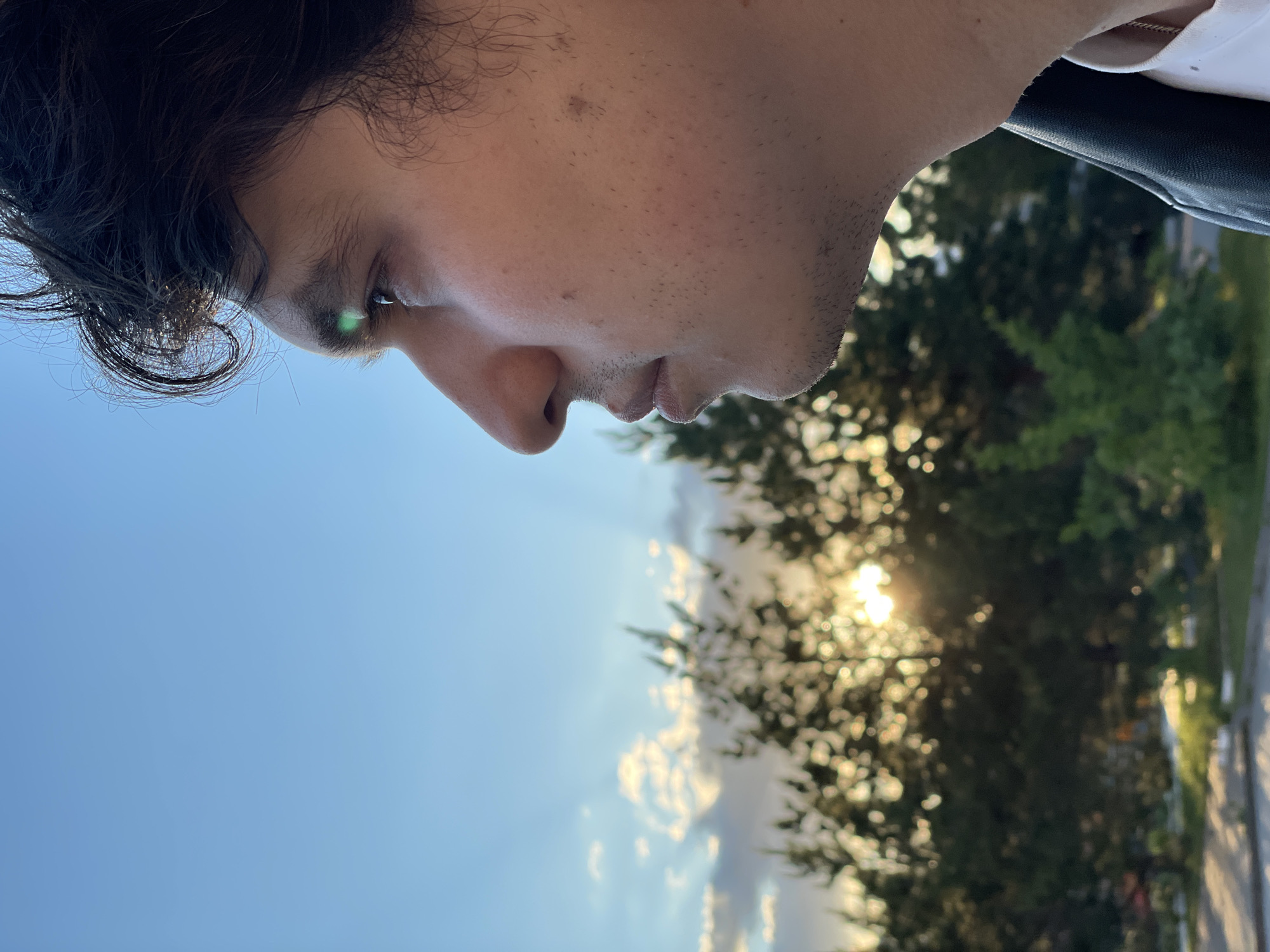 The image displays a close-up, profile shot of a person, likely male, oriented as if looking to the right. The individual has dark, curly hair visible on the top and left side of their head. Their skin appears light-toned, with visible stubble on the cheek and chin, and a few small moles or freckles on the cheek. One eye is prominent, reflecting a noticeable green lens flare. The person's gaze is directed forward and slightly downward, conveying a contemplative or observant mood.
A white collar or shirt is visible on the far right edge of the frame, adjacent to a dark, possibly leather-like material, which could be part of an outfit or a car interior.
The background reveals an outdoor setting under a clear blue sky with some scattered clouds. The sun is low in the sky, positioned behind and to the right of the person, casting a warm, golden light that filters through dense green foliage, likely trees. The background is heavily blurred with a strong bokeh effect, especially around the illuminated leaves and the bright sun. The low angle and warm light from the sun suggest the time of day is either sunrise or sunset. No visible text is present in the image.