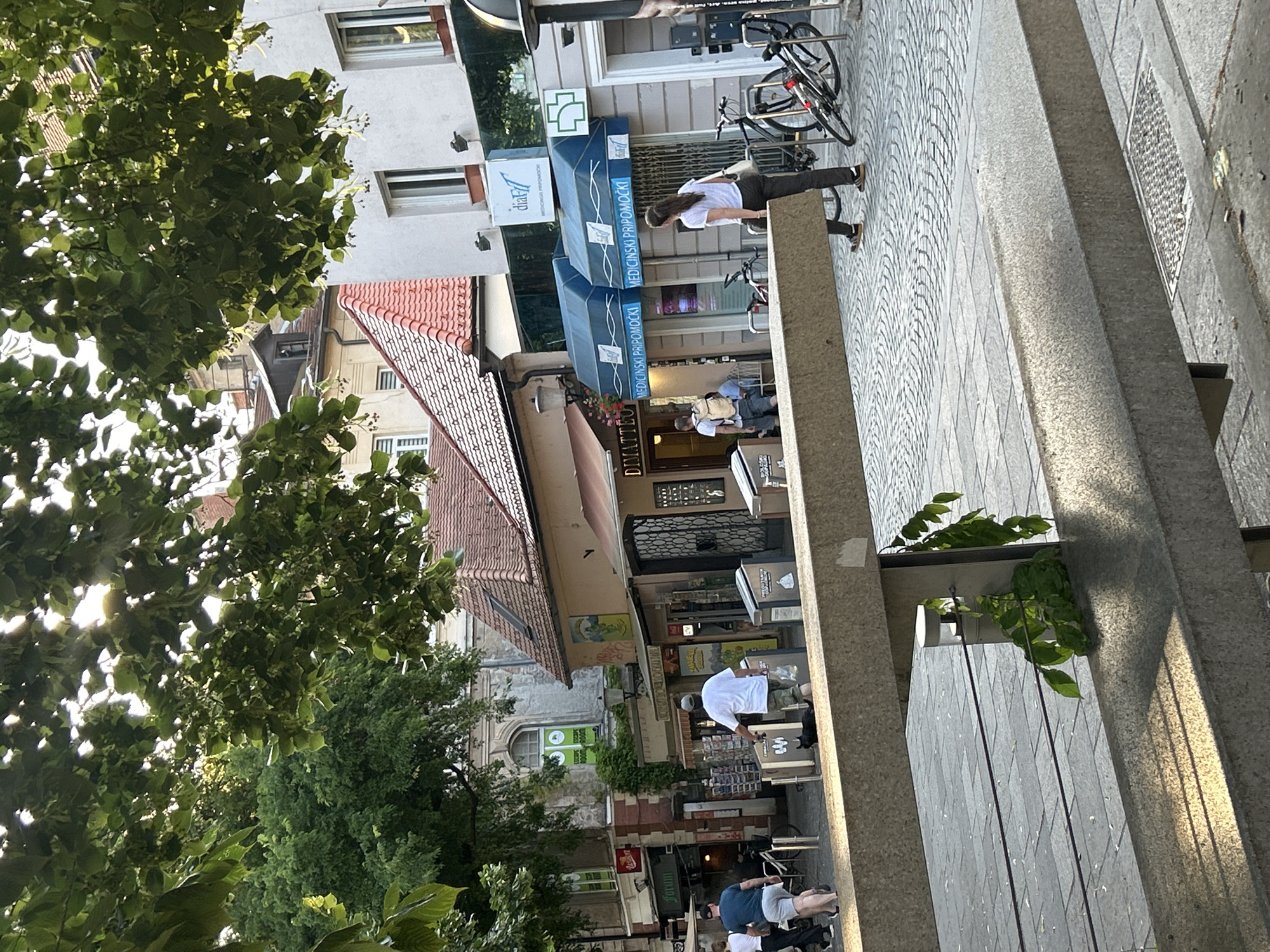 The image shows a street scene with several buildings and people. In the foreground, a stone bench is visible with a plant growing from a metal post behind it. Beyond the bench, a cobblestone street leads to buildings with awnings and shop fronts. Several people are walking on the street, including a woman in a white shirt and dark pants, a man with a backpack, and a man in a white shirt and camouflage shorts. A black cat is also present on the street. In the background, trees with green leaves overhang the scene, and the sun is shining through them, creating dappled light. The buildings appear to be a mix of old and new, with some featuring tiled roofs and others modern facades. Some of the shop signs are in a foreign language.