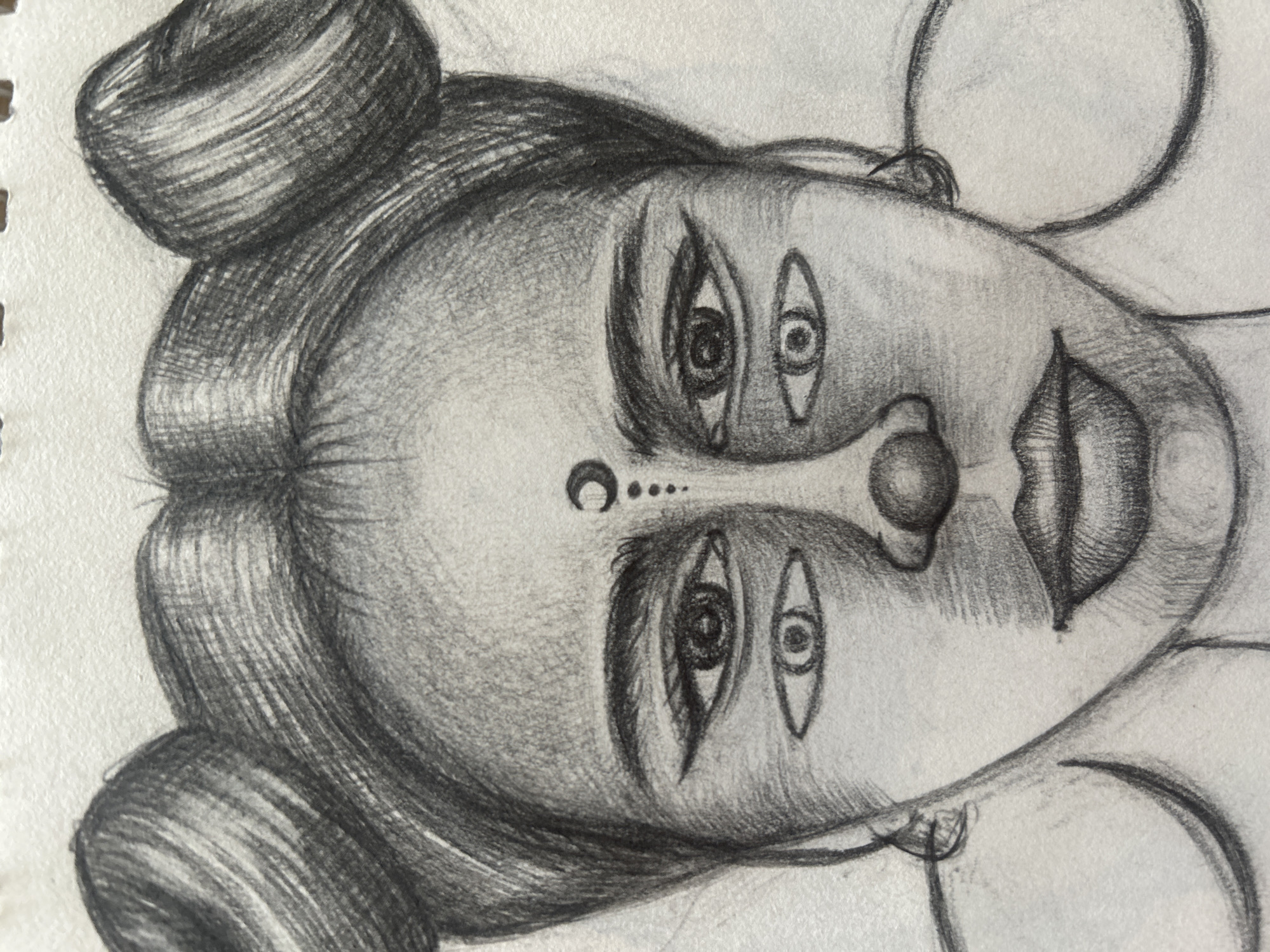 The image is a black and white pencil sketch of a face with multiple eyes. The subject has dark hair styled in two buns on top of her head. Her forehead is adorned with a crescent moon symbol and a series of dots. She has large eyes, with an additional set of eyes drawn below each main eye. Her nose is rounded, and her lips are full. She is wearing large hoop earrings. The background is blank, suggesting the sketch is on paper. There is no discernible location in Moscow, Russia, as it is an artistic depiction.