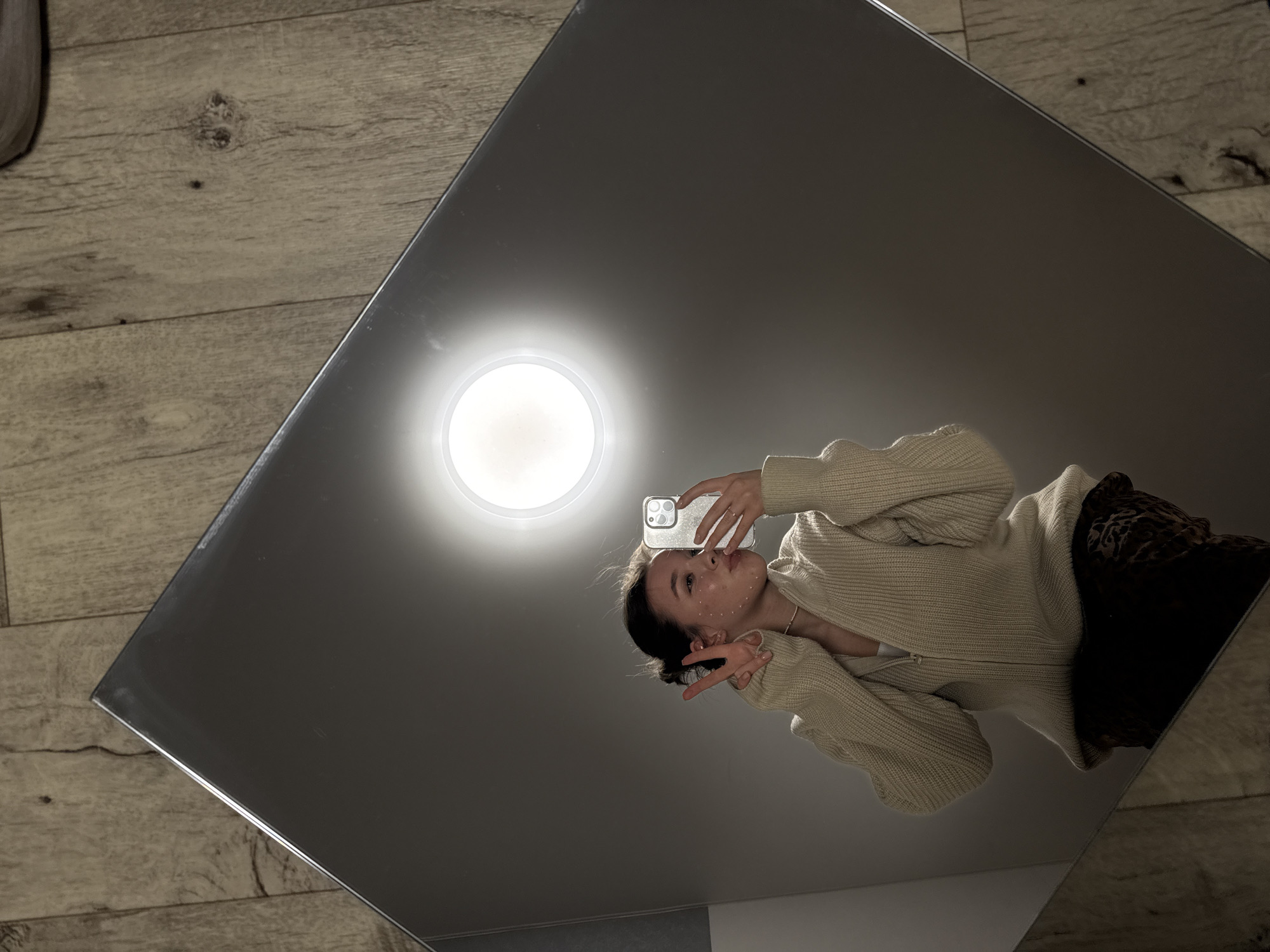 A young woman is reflected in a square mirror placed on a wooden floor with light wood-grain planks. The woman is facing the mirror, holding a mobile phone up to take a selfie, her face is visible in the reflection. She wears a cream-colored, knitted cardigan. A light fixture is visible in the mirror above her head. A small portion of a wall is visible in the corner of the mirror. There is no indication of the location in the city of Noviy, Russia.