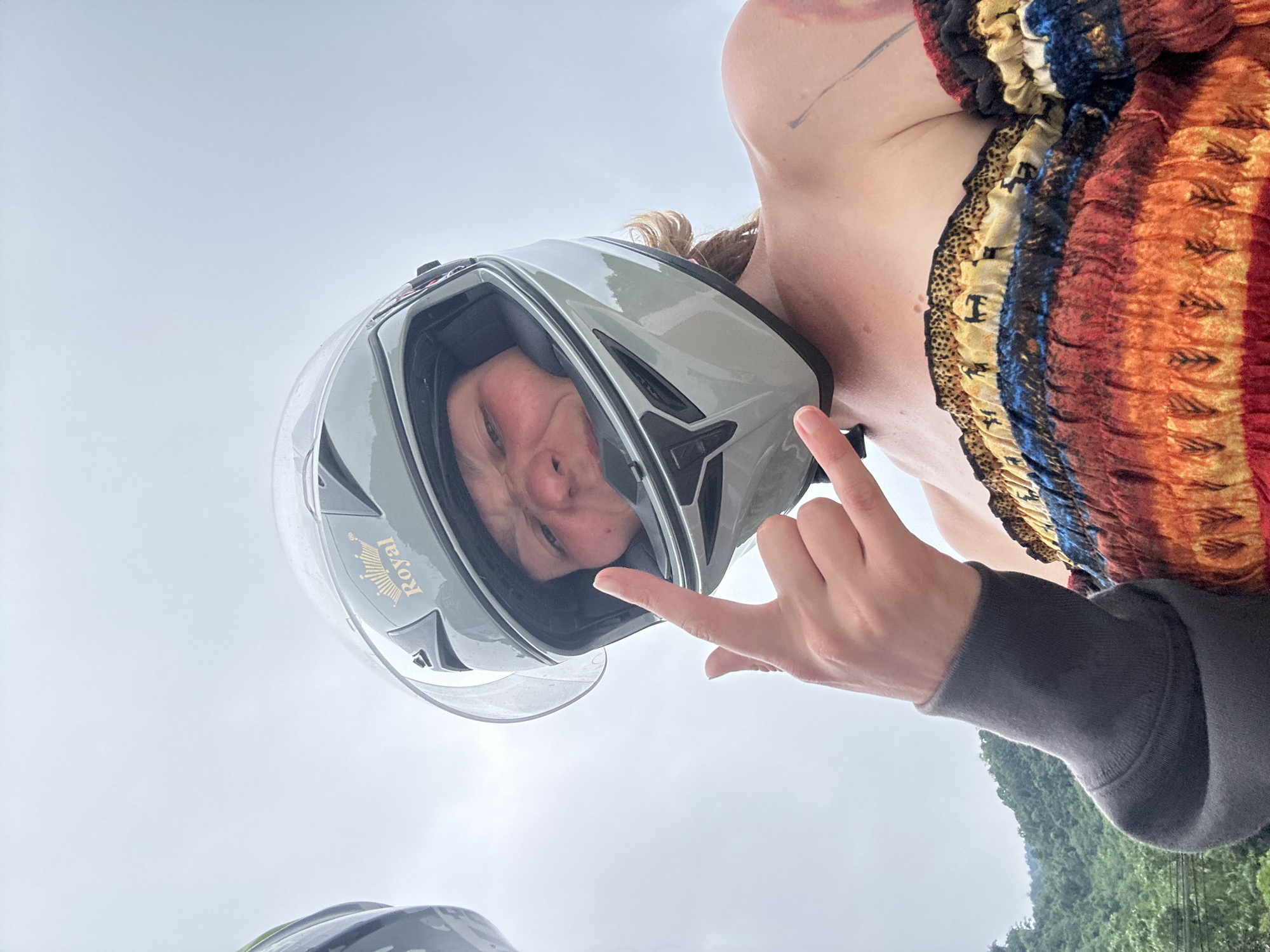 A low-angle, full-body shot shows a person wearing a grey motorcycle helmet and a colorful off-the-shoulder dress. The person is making a rock-and-roll hand gesture with their right hand, with their left arm visible wearing a dark gray sleeve. The background shows a cloudy sky and lush green hills, suggesting a rural or mountainous area. The specific location is not discernible from the image itself; however, it is described as being in Đồng Bảng, Vietnam.
