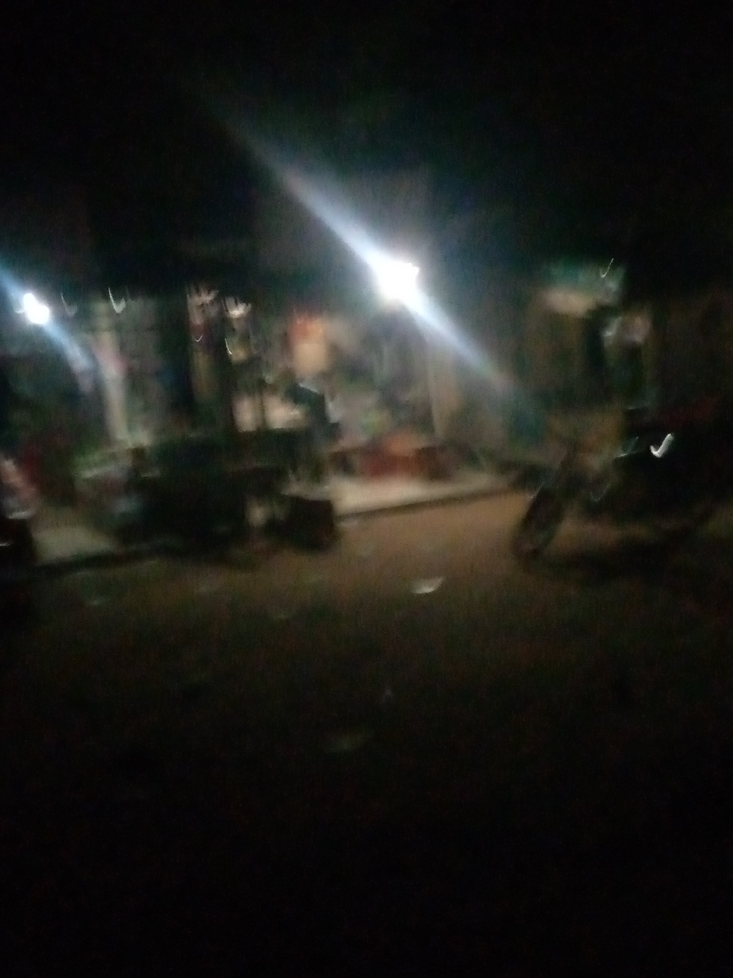 The image depicts a dimly lit outdoor scene, likely at night, in Jalingo, Nigeria. The focus is on a street or pathway with some structures in the background, possibly shops or residences. A bright light source, possibly a streetlamp or the headlights of a vehicle, illuminates the scene from the left, casting long streaks of light across the frame. In the mid-ground, several objects are present, including what appears to be a motorcycle or bicycle, with its form blurred due to the low light and possible movement. The ground is dark and appears to be unpaved. The overall impression is of a busy, yet obscured, urban environment.