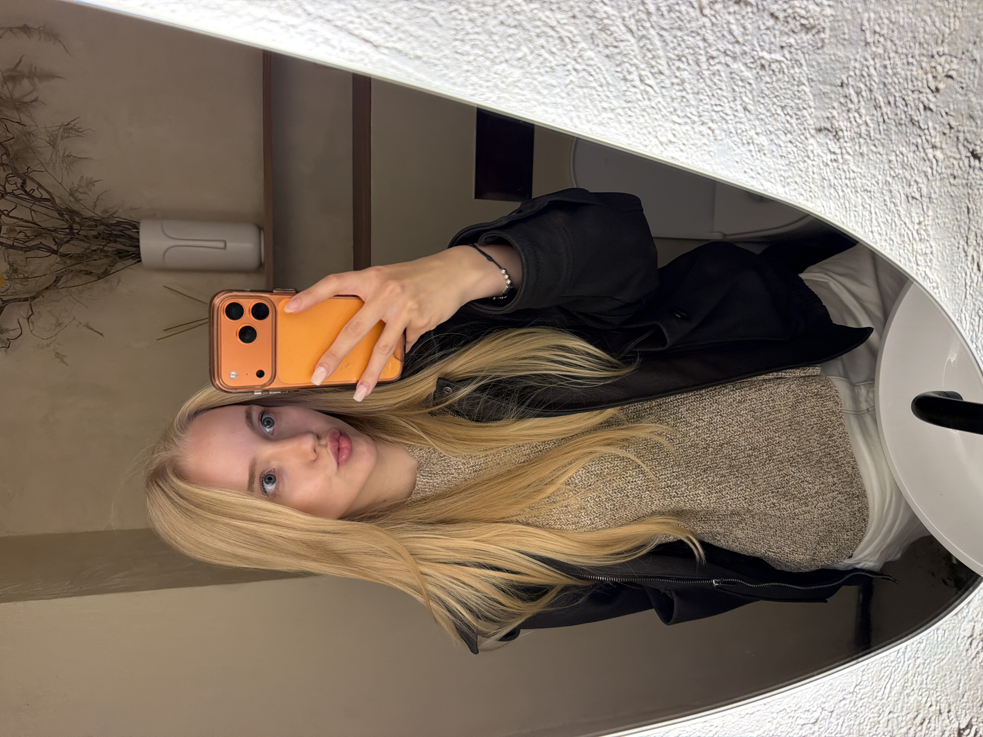 A young, fair-skinned female with long, straight, light blonde hair and blue eyes is captured in a mirror selfie. She holds an orange-cased smartphone with a visible triple camera array in her right hand, angling it to photograph her reflection. Her lips are puckered, forming a "duck face" expression. She wears a black jacket over a light brown or beige textured knit sweater. A simple black cord bracelet adorned with several small silver beads is present on her left wrist. Manicured, light-colored fingernails are visible on her right hand. The mirror, framed by a textured, light-colored border, reflects a bathroom or lounge setting. A white ceramic sink with a black faucet is visible in the lower part of the reflection. Behind the female, a light taupe wall is visible, featuring a light wooden shelf. On the shelf, a white vase with a minimalist face design holds dried, light brown foliage, and several thin dark diffuser sticks are placed to the left of the vase. The lighting appears uniform and indirect. The location is an indoor facility in the city of Unknown, Unknown.