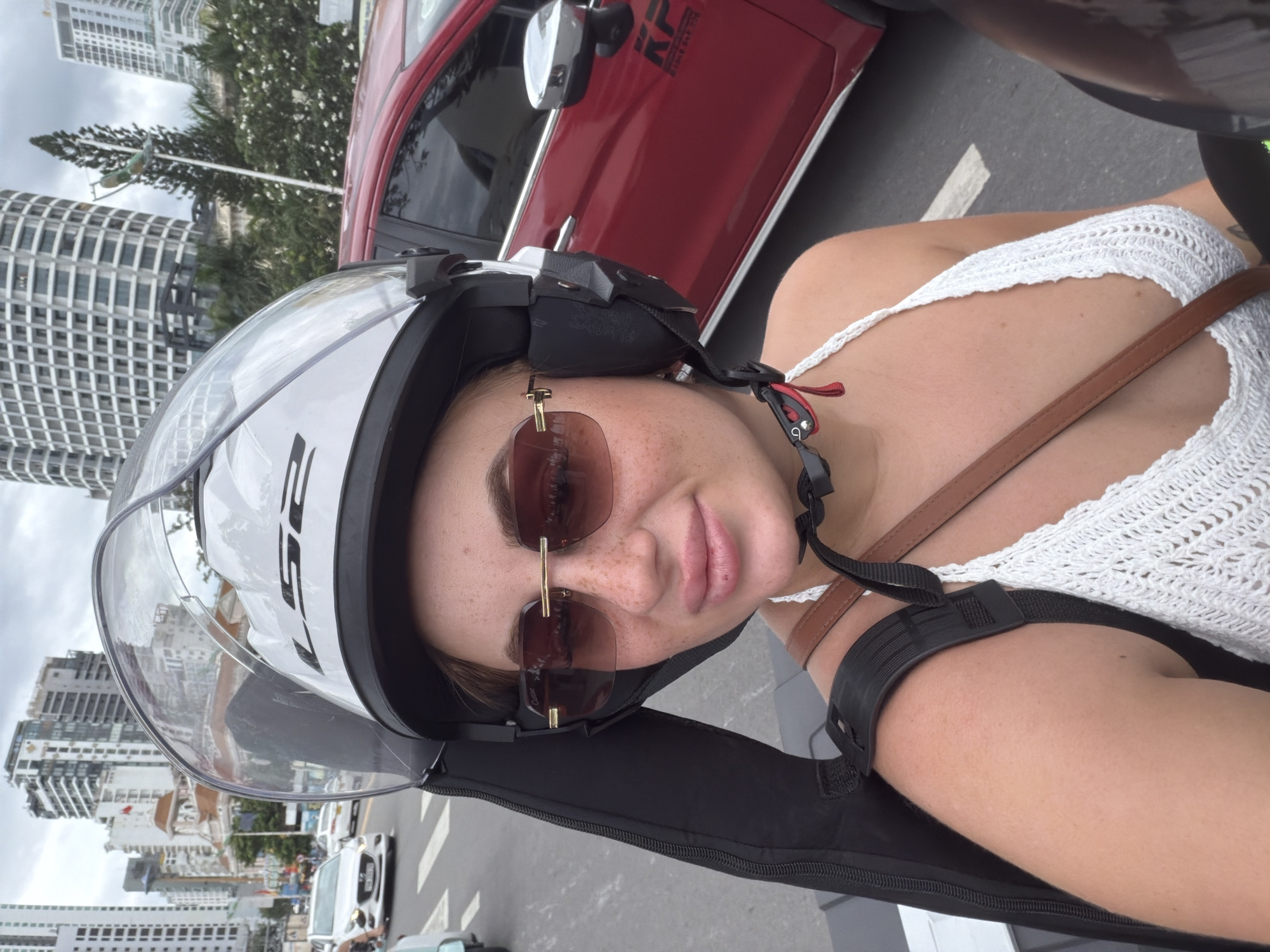 A woman wearing a white crocheted top and a white helmet with a clear visor is riding a motorbike in Nha Trang, Vietnam. She has on large, square, brown-tinted sunglasses. The woman is looking directly at the camera. She has freckles across her nose and cheeks. A dark case is strapped to her back. A red car is visible to her right. In the background, tall buildings line the street, indicating an urban setting. The sky is overcast.