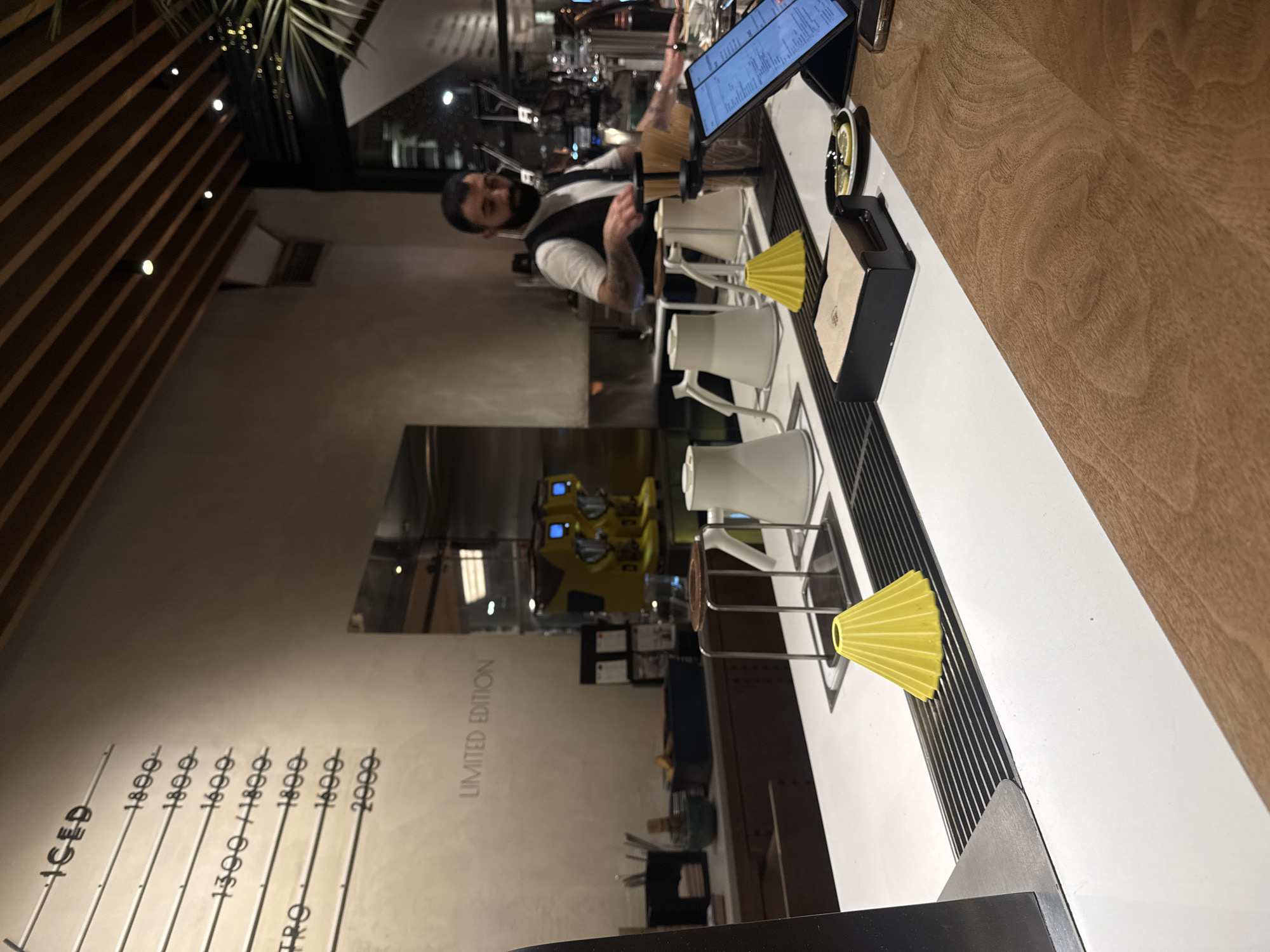 The image depicts the interior of a modern coffee shop or cafe, likely located in Yerevan, Armenia, based on the monetary values (e.g., 1800, 1600) displayed on the menu, which correspond to Armenian Dram.
In the foreground, a light brown wooden counter transitions to a white countertop equipped with a black metal grate. Two distinct yellow, pleated, conical objects rest on this grate, positioned in front of two white pour-over kettles and two metal pour-over stands designed for coffee preparation. To the right, a black napkin holder containing brown napkins is visible next to a small black dish holding lemon slices. A black tablet on a stand, displaying digital text, is positioned further right, alongside a smartphone.
In the midground, behind the white counter, a male barista with dark hair and a beard is present. He wears a white short-sleeved shirt, a dark vest, and a dark face mask, with visible tattoos on his right arm. He appears engaged in preparing beverages, his hands near various pieces of bar equipment.
The background features a light-colored wall with a menu displaying items such as "ICED" and "LIMITED EDITION," alongside several numeric values (e.g., 1800, 1600, 1300/1800, 2000). Two prominent yellow espresso machines with blue screens are visible on a counter behind the barista. Other bar essentials, including containers and shelving, are arranged on a dark counter against the wall. A large mirror reflects parts of the interior, and through a window or glass partition in the far background, blurred lights suggest an exterior nighttime street scene. The ceiling is characterized by horizontal wooden slats with embedded circular lights. Green foliage from a plant is partially visible in the upper right corner.
