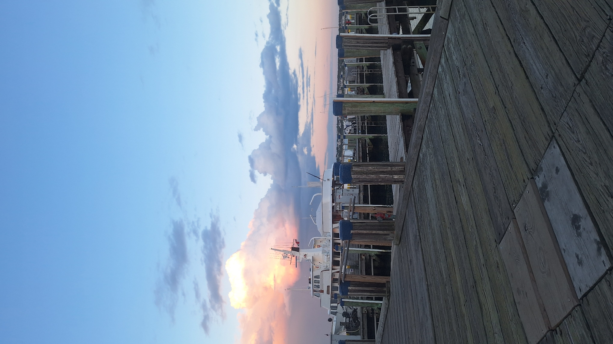 The image shows a view from a wooden dock looking out at a harbor. A white ferry boat is docked on the left, with other boats and docks visible in the background. The sky is a mix of blue and pink hues, with large clouds illuminated by the setting sun. The wooden planks of the dock are worn and weathered, suggesting a seaside location. The scene is quiet and peaceful, with the gentle lapping of water against the boats and docks.