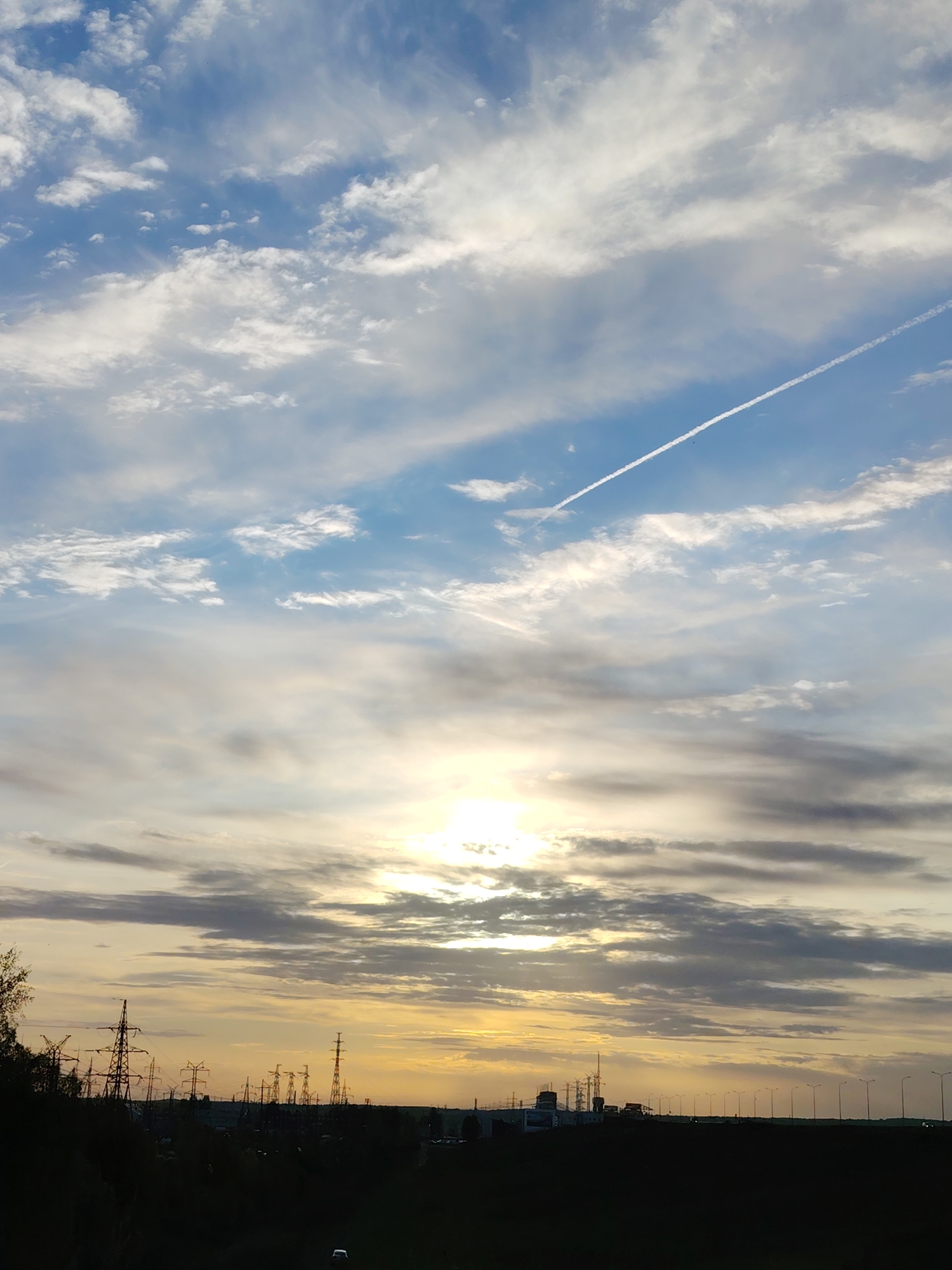The image depicts a landscape at sunset in Chaikovsky, Russia. Overhead, the sky is a mix of blue and grey clouds, with a visible contrail from an aircraft cutting across the upper right portion of the sky. The sun, positioned lower in the frame, is partially obscured by a layer of clouds, creating a diffused, golden glow.
The lower portion of the image showcases a silhouette of trees and infrastructure. Several power lines and transmission towers are visible against the sky. In the distance, a row of street lights are evenly spaced along a road. The foreground is a dark, undefined mass of land. A single vehicle is present on the road.