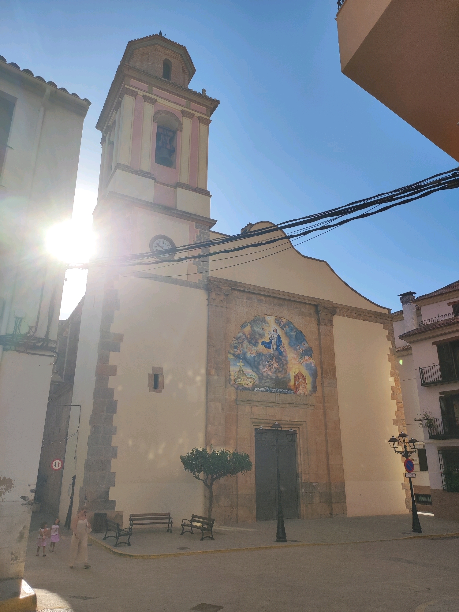 The photograph captures a view of a church in the town of Suera, Spain, under a clear blue sky. The sun is bright, casting lens flares on the left side of the image. The church features a bell tower with a clock face and a pitched roof. The facade is painted a light color, with a large, colorful tiled mural above the entrance doors depicting a religious scene. To the right of the church, a multi-story building with balconies is visible. In front of the church, a paved area has benches and a small tree. Two young girls in pink dresses are walking in the foreground to the left, accompanied by an adult woman in a light-colored outfit. A lamppost stands on the right side of the paved area. Power lines are strung across the upper part of the image.