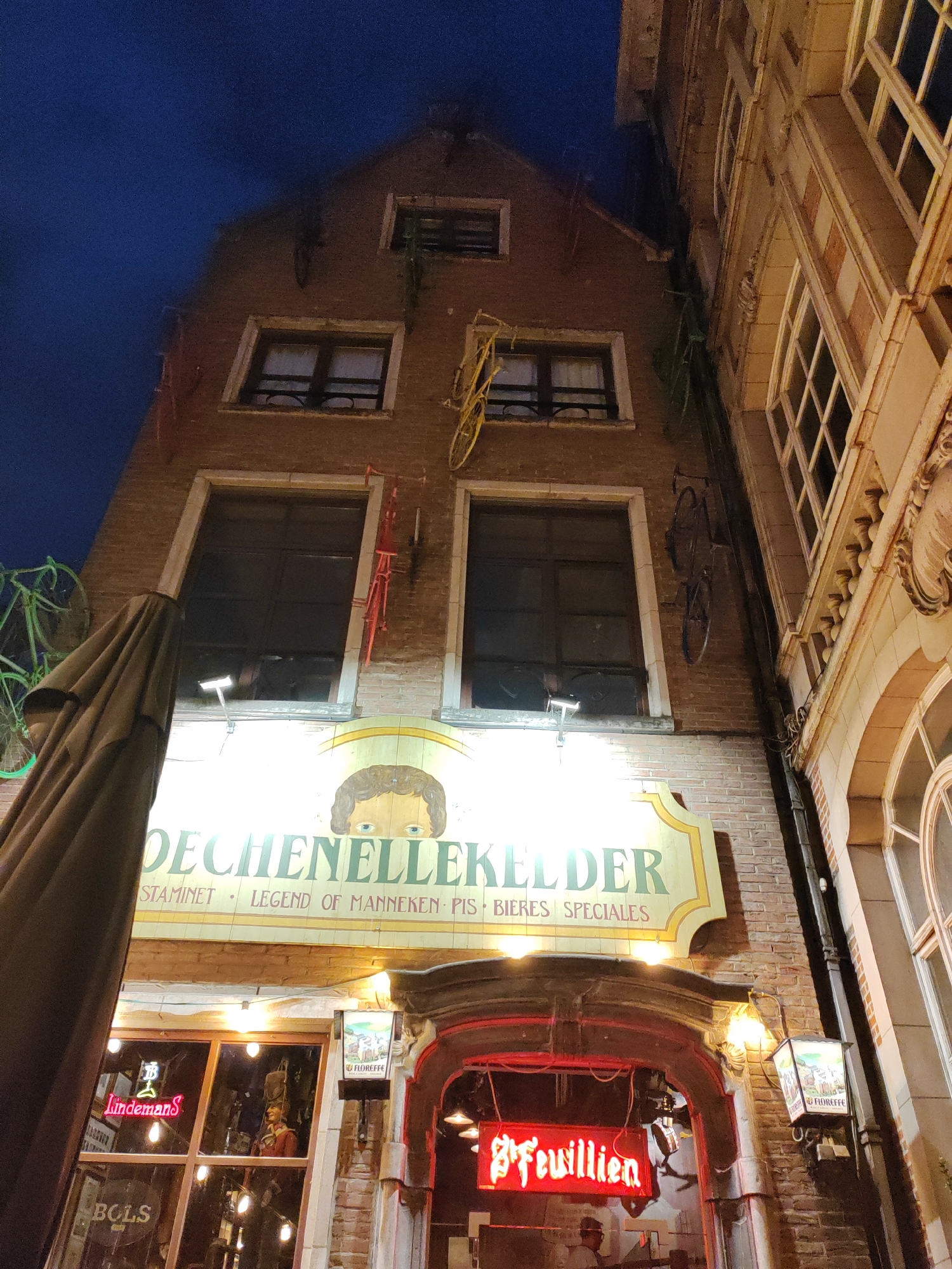 The image shows a night-time exterior view of a narrow brick building in Brussels, Belgium. The building has several windows and is decorated with bicycles hanging from the exterior walls. A large sign with yellow and green lettering reads "DECHENELLEKELDER" above a smaller text that appears to say "STAMINET LEGEND OF MANNEKEN PIS BIERES SPECIALES".
Below the sign, the entrance to the establishment is visible, with a red neon sign that reads "FEUILLIEN". To the left of the entrance, through a window, a neon sign for "Lindemans" can be seen, along with bottles and a figurine. Another sign for "BOLS" is also visible through the window. To the right of the entrance, a lantern with an illustration is mounted on the wall. The sky above is dark blue. The adjacent building to the right has a lighter-colored facade with numerous windows. The overall scene suggests a bar or restaurant.