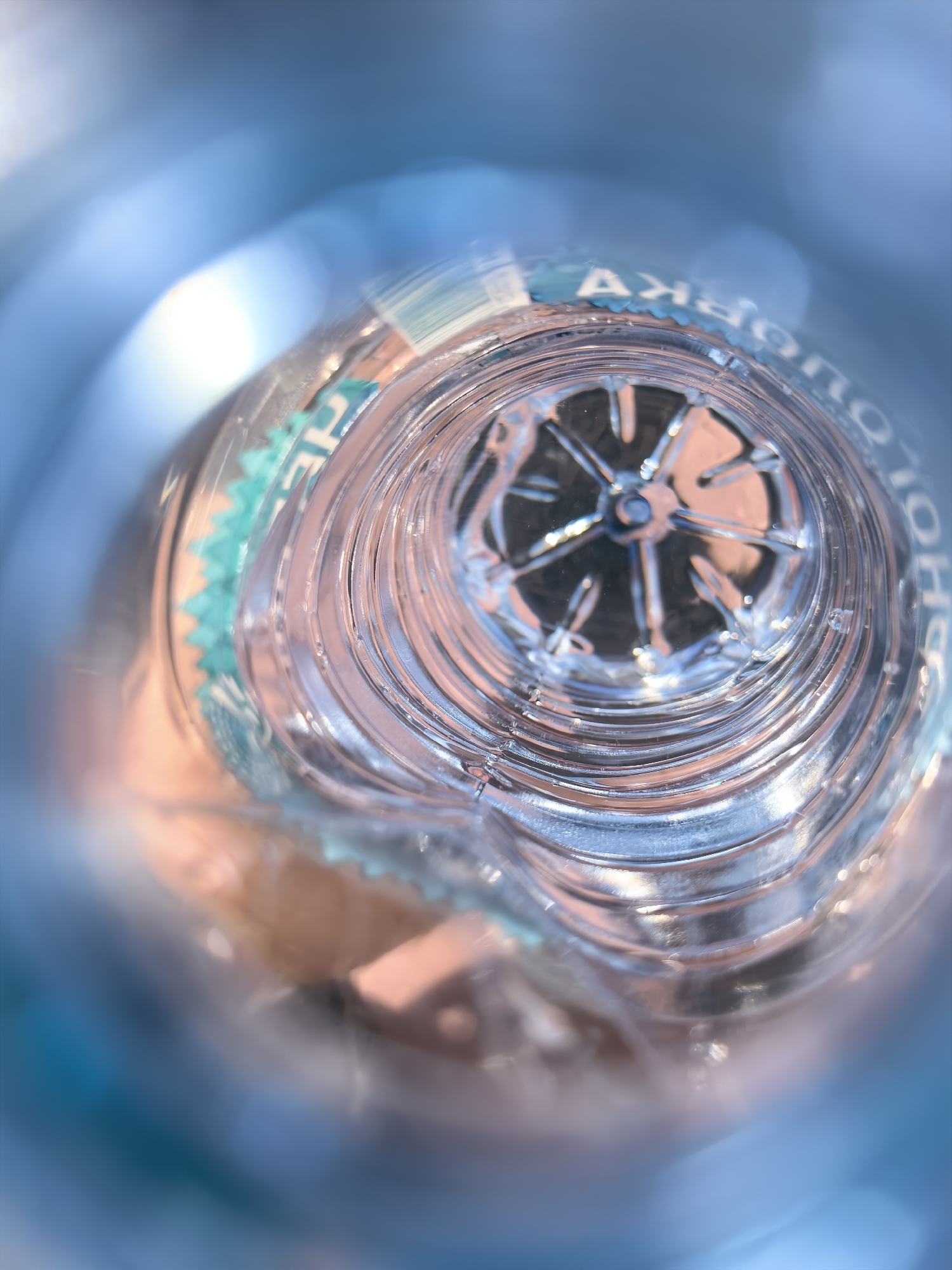 An extreme close-up shot looks down the opening of a clear plastic water bottle. The bottle's neck is ribbed, and the bottom of the bottle has a star-shaped pattern. The background is blurred blue and pink hues, suggesting sunlight reflecting off surfaces. No people or specific city locations are visible.