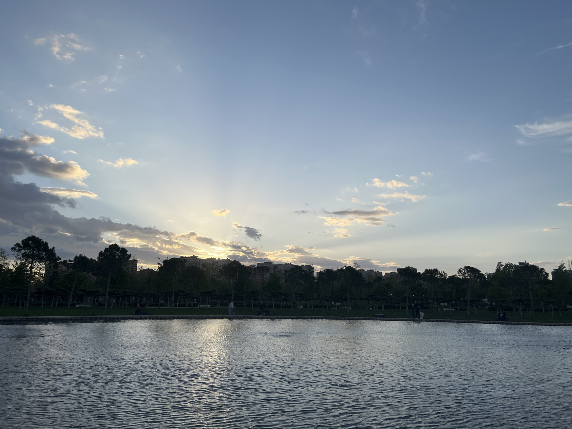 The image shows a park scene at dusk. A large body of water, possibly a lake or pond, dominates the foreground. The water's surface is rippled. In the middle ground, there's a strip of green space with trees. Behind the trees, buildings are visible. The sky is a light blue with scattered clouds. The sun is setting, casting light rays through the clouds. People are visible near the green space, engaging in activities like walking or sitting. The location is likely a park in a city; based on the prompt, that would be Krasnodar, Russia.