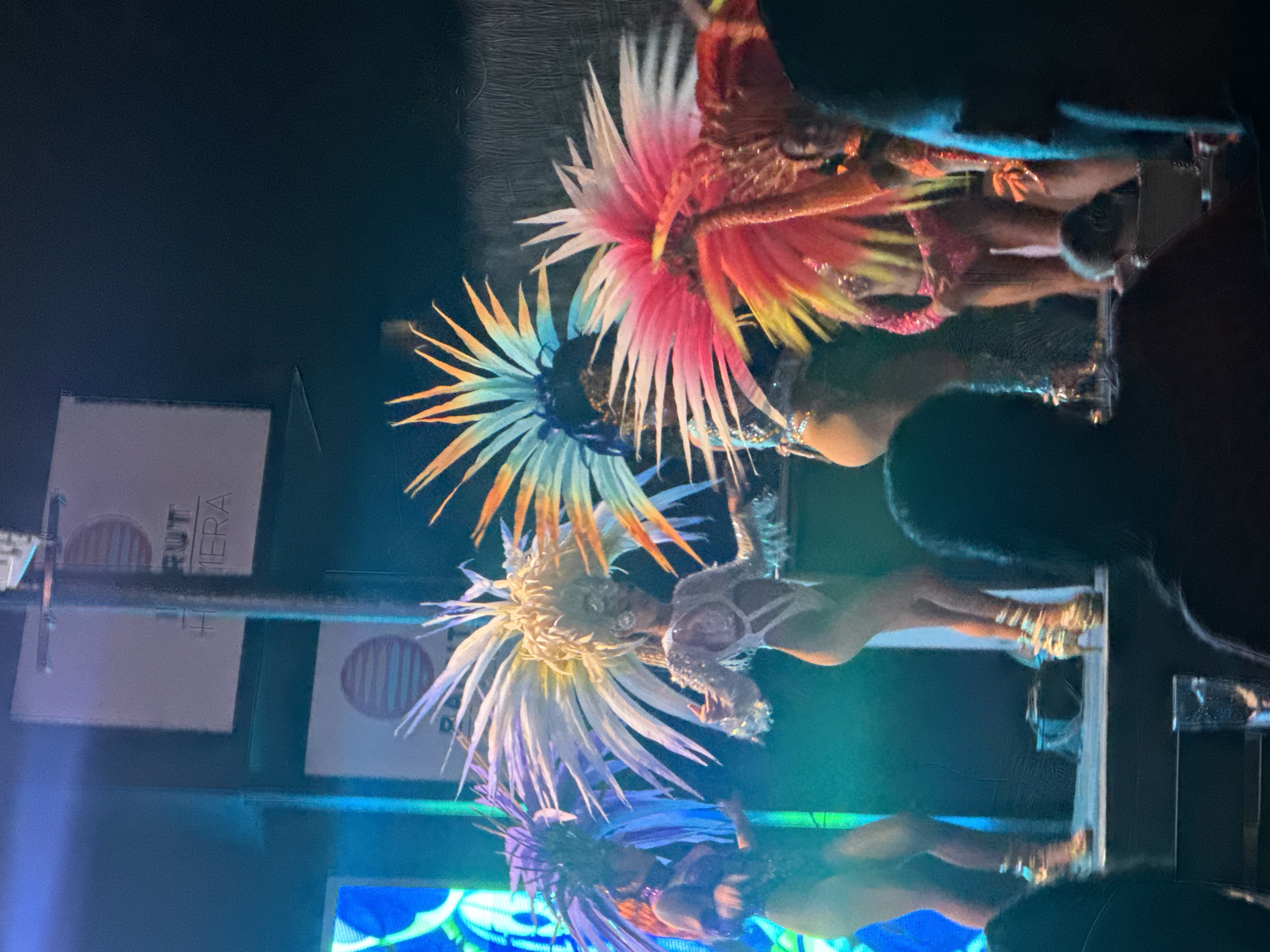 The image captures a vibrant performance on a stage in Manavgat, Turkey. In the foreground, the silhouettes of audience members obscure the lower portion of the scene. The stage is lit with blue and green lighting, illuminating three performers dressed in elaborate, colorful costumes. They are wearing large, feathered headdresses and sequined outfits, suggesting a cabaret or showgirl performance. The performer on the far left wears a purple headdress and a dark, shimmering outfit. The central performer sports a large, white and yellow headdress and a light-colored, cut-out costume with sparkly accents. The performer to the right has a bright pink and yellow headdress and a pink costume. Behind the performers, a sign with a circular logo and text is visible, though partially obscured. The overall atmosphere appears energetic and festive, typical of a nightlife entertainment venue.