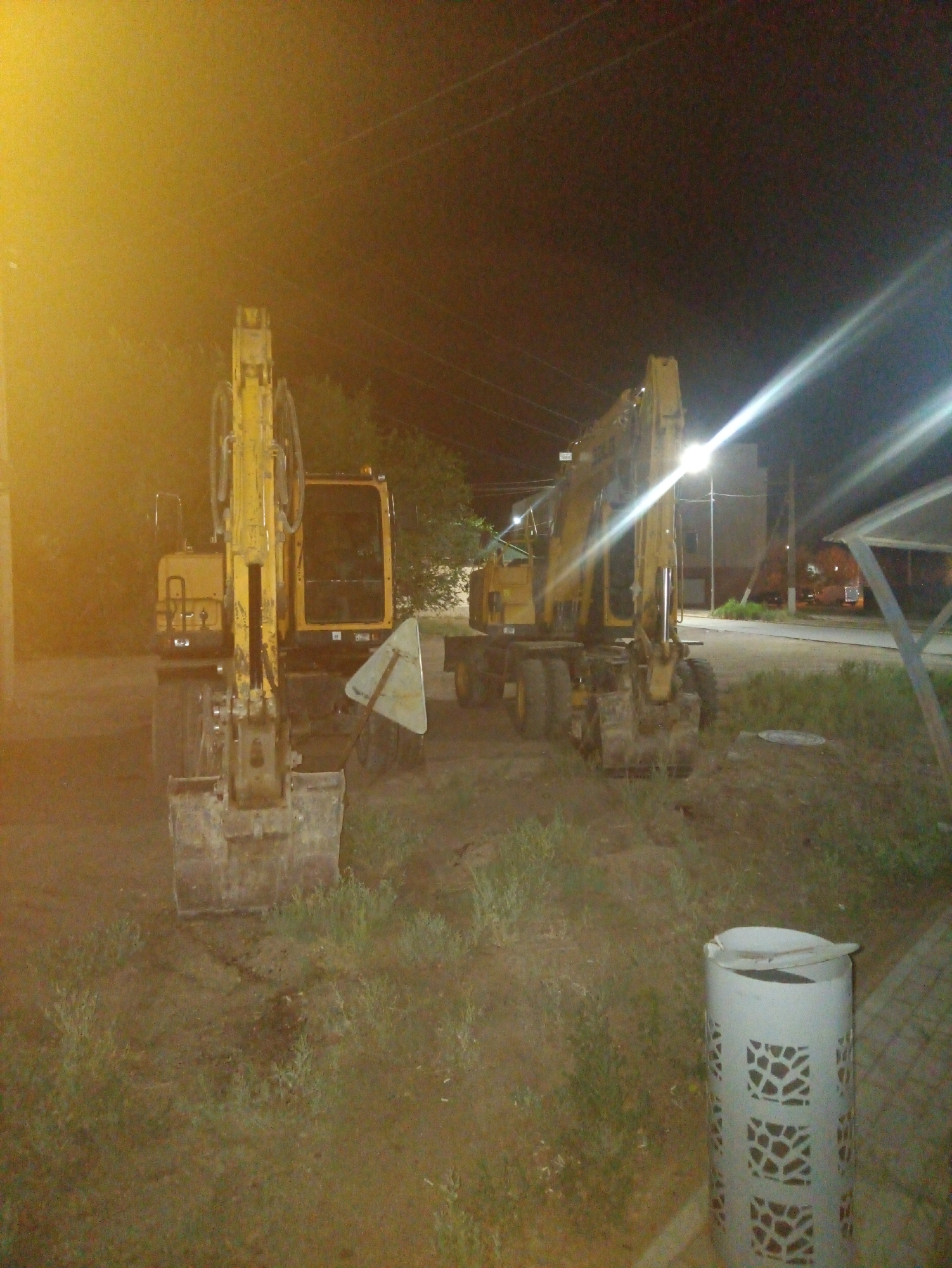 The image depicts two yellow excavators parked on an unpaved area at night. The left excavator is closer to the camera and is facing forward. The right excavator is further away and facing to the right. The scene is illuminated by artificial lights, creating bright glare effects. In the foreground, there is a white cylindrical trash can with a decorative pattern. The background shows a street with a sidewalk, some buildings, and utility poles. There are trees and vegetation visible on the sides. The ground appears to be dirt and grass. The overall setting suggests an outdoor industrial or construction area, possibly in the city of Balqaş, Kazakhstan, given the context. There are no people visible in the image.