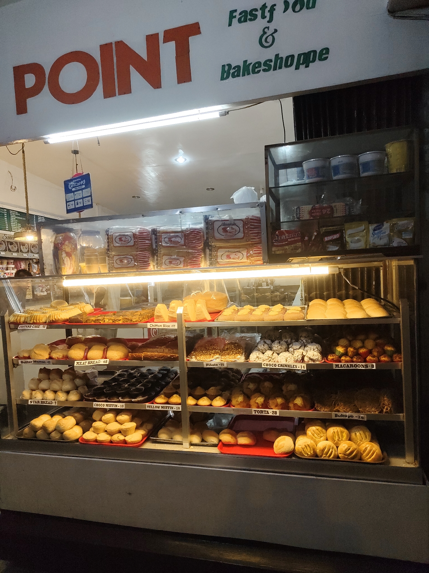 This media file shows the exterior of a bakeshop named "POINT Fastf & Bakeshoppe" in Davao City, Philippines. The sign "POINT" is prominently displayed in large orange letters above the entrance. The bakeshop has a display case filled with a variety of baked goods, including cakes, breads, muffins, and cookies. Some of the items are labeled with their names and prices, such as "CUSTARD CAKE - 48", "Butter Mamon - 17", "MEAT BREAD - 48", "CHOCO MUFFIN - 10", and "MACAROONS - 8". There are also packaged goods like "QuickChekcha" and "Kopiko" visible on a shelf. A blue sign for "GCash Po" is hanging near the entrance. The lighting suggests it might be evening or indoors. The scene depicts a commercial establishment ready to serve customers.