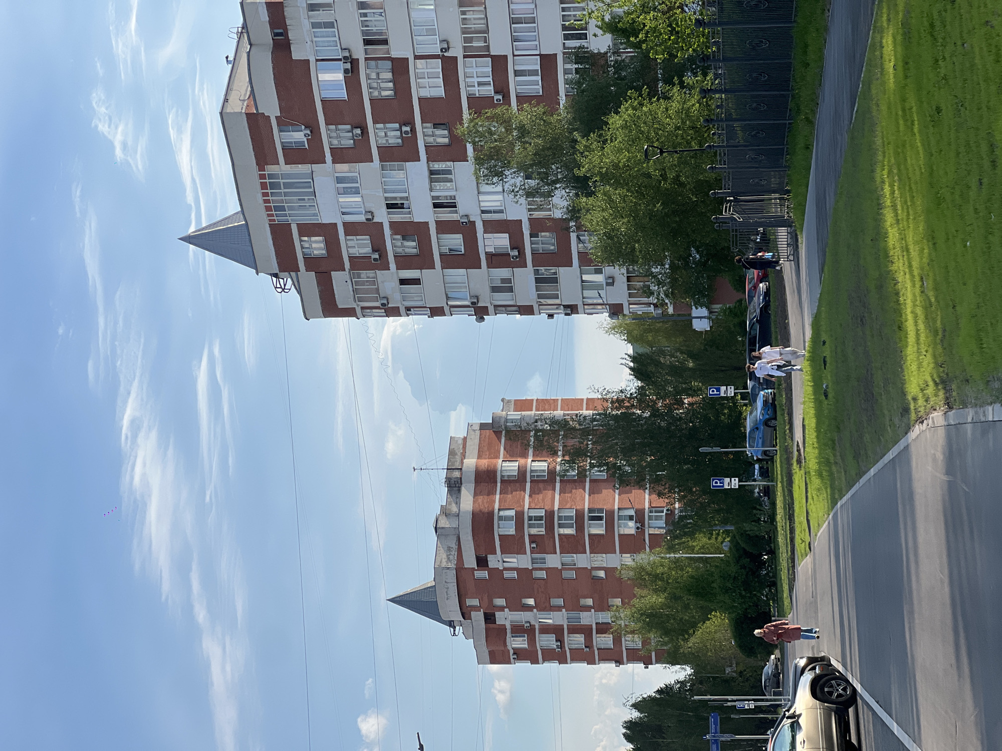 A street scene in Moscow, Russia is presented from a ground-level perspective. Two tall buildings with red and white facades stand in the background, with a pointed roof on the top floor of each. Green trees grow along the road, next to a fence. Several people are walking on the sidewalk, and parked cars are seen alongside the road. Street signs with the letter "P" can also be seen. The sky is blue with scattered clouds.