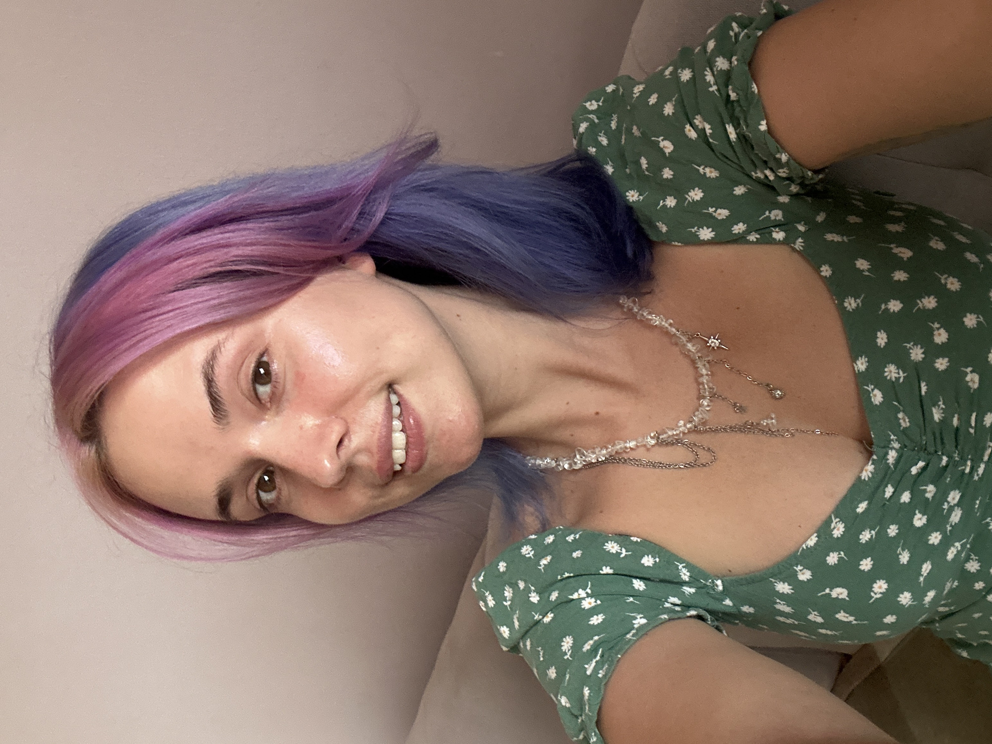 A woman poses for a selfie, her head tilted slightly to the side. She has pink and blue hair. She wears a green dress with white floral prints and a beaded necklace. She is smiling. The background is out of focus and indistinct, but appears to be an indoor setting. There is no discernible information to place the location in Gijón/Xixón, Spain.