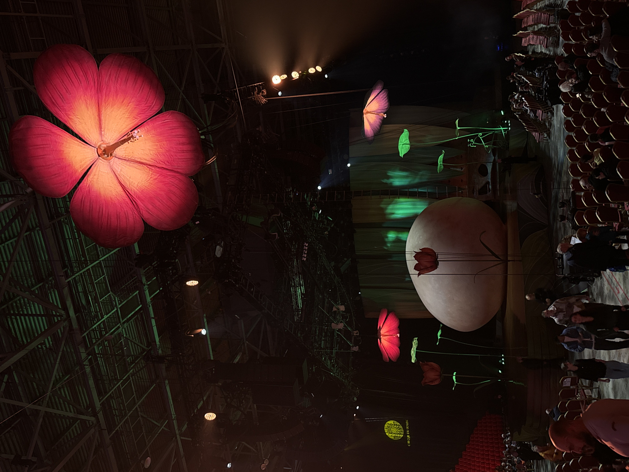 The image shows an indoor stage setup for a theatrical performance, possibly Cirque du Soleil. The scene is bathed in dramatic lighting. In the foreground, a large, red, illuminated flower prop dominates the left side of the frame. Further back, a giant white egg-shaped prop sits in the middle of the stage, with several smaller, red tulip-like structures attached to it. A butterfly prop is suspended in the air. Seating is arranged to the right. Several people are visible standing and milling about in the seating area. The scene takes place within a large industrial structure, based on the metal support beams and rigging visible in the background. The image does not provide enough information to determine if it's in Sofia, Bulgaria.