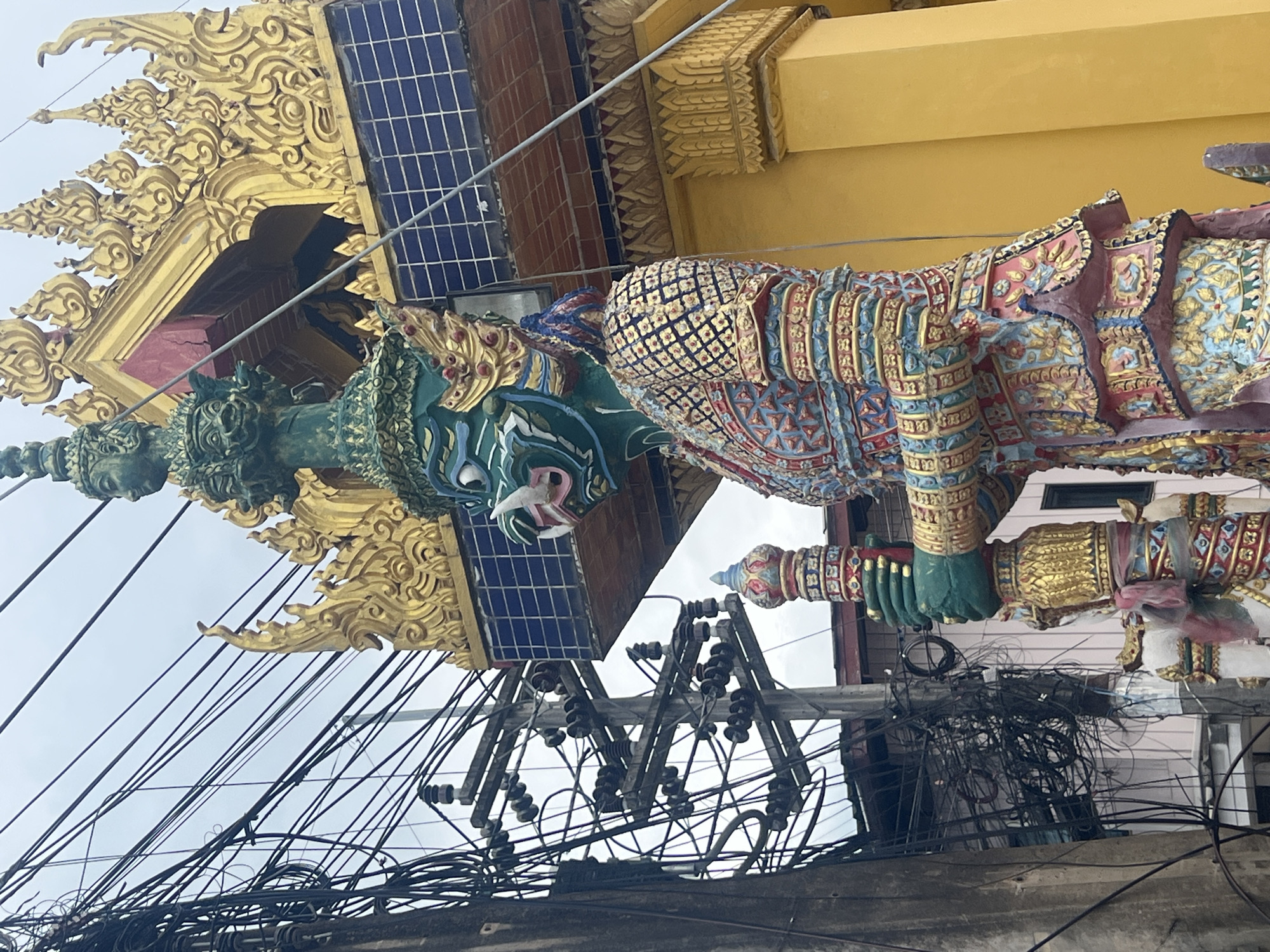 An exterior, medium, low-angle shot shows a towering, ornate, green-faced statue of a mythical guardian, likely a yaksha or demon, standing in front of a yellow building with a gold, intricately carved roof. The statue is highly detailed, adorned with colorful, patterned armor in shades of red, blue, gold, and pink. It holds a cylindrical object, possibly a staff or weapon, in its green hands. To the left of the statue, a dense tangle of black electrical wires crisscrosses in front of a grey concrete pole and a lighter colored building with pink siding. The sky above is overcast and pale. The scene is a street view with a juxtaposition of spiritual art and urban infrastructure.