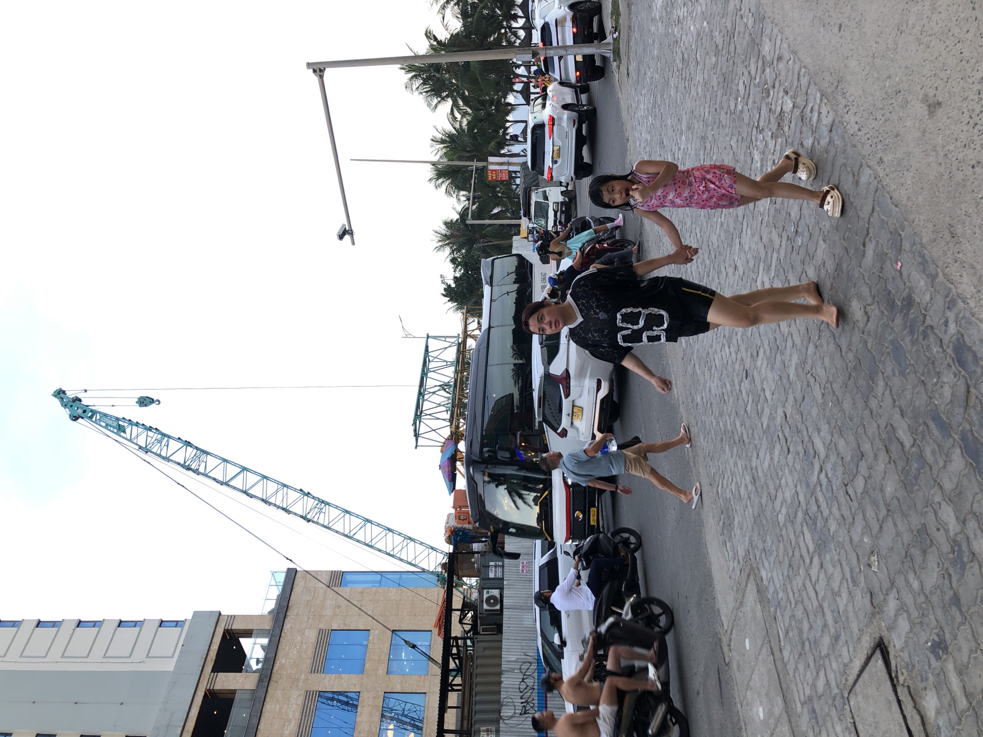 In the city of Đà Nẵng, Vietnam, a street scene unfolds with a mix of construction and daily life. A tall, blue construction crane dominates the background, its arm extended over a building under construction. In the foreground, a woman and a young girl walk hand-in-hand on a cobblestone sidewalk. The woman wears a black athletic jersey with the number "69" on it and black shorts, while the girl is dressed in a pink patterned swimsuit.
To their left, several people are on motorcycles, with two men in the foreground riding a scooter. Behind them, a white SUV and a black bus are visible in traffic, along with other vehicles. Palm trees line the street, suggesting a tropical coastal location. The sky is overcast, with a pale white hue. The overall impression is a busy urban environment with ongoing development.