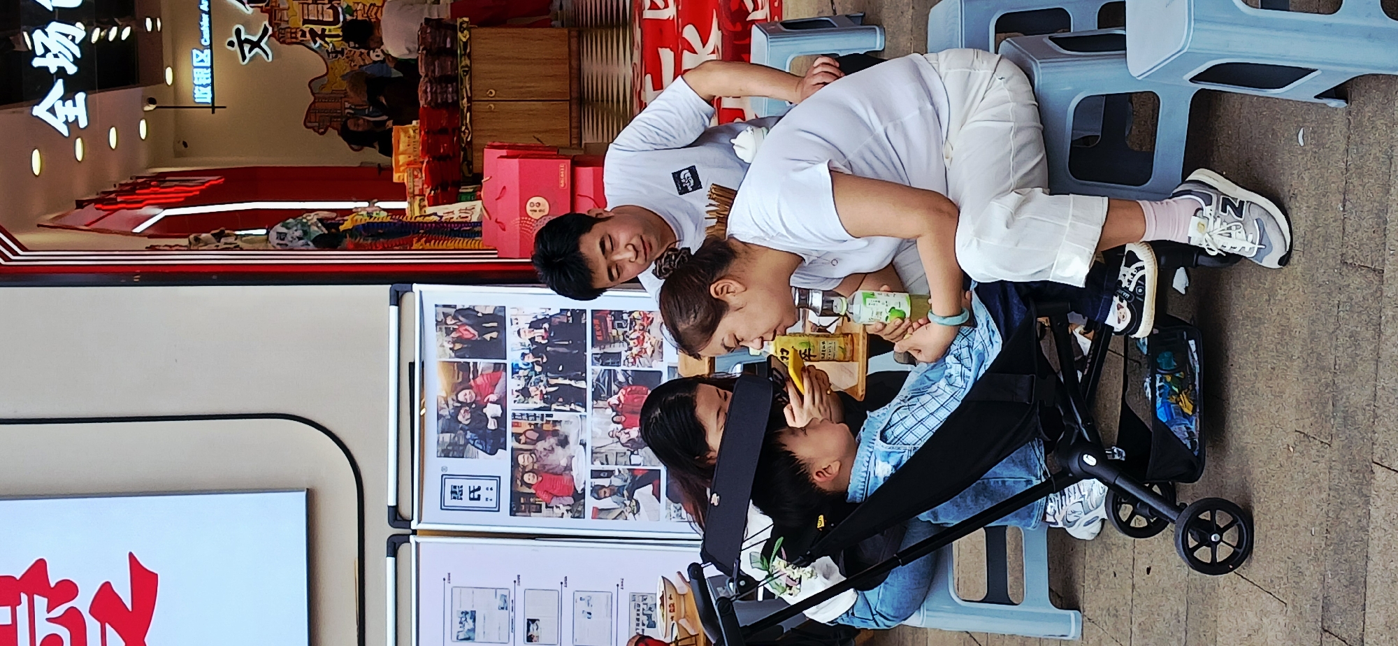 The image, taken outdoors in Yuzhong District, China, shows two adults and a child interacting beside a stroller. The adults, a man and woman both wearing white shirts, are leaning over the stroller where a child sits. The woman is feeding the child from a bottle. One of the adults holds a phone, and there are two other bottled drinks visible. They are positioned next to a sign with photographs and writing in Chinese. The background includes the exterior of a building with Chinese signage, suggesting a commercial district. There are blue plastic stools nearby, and the ground appears to be a sidewalk.