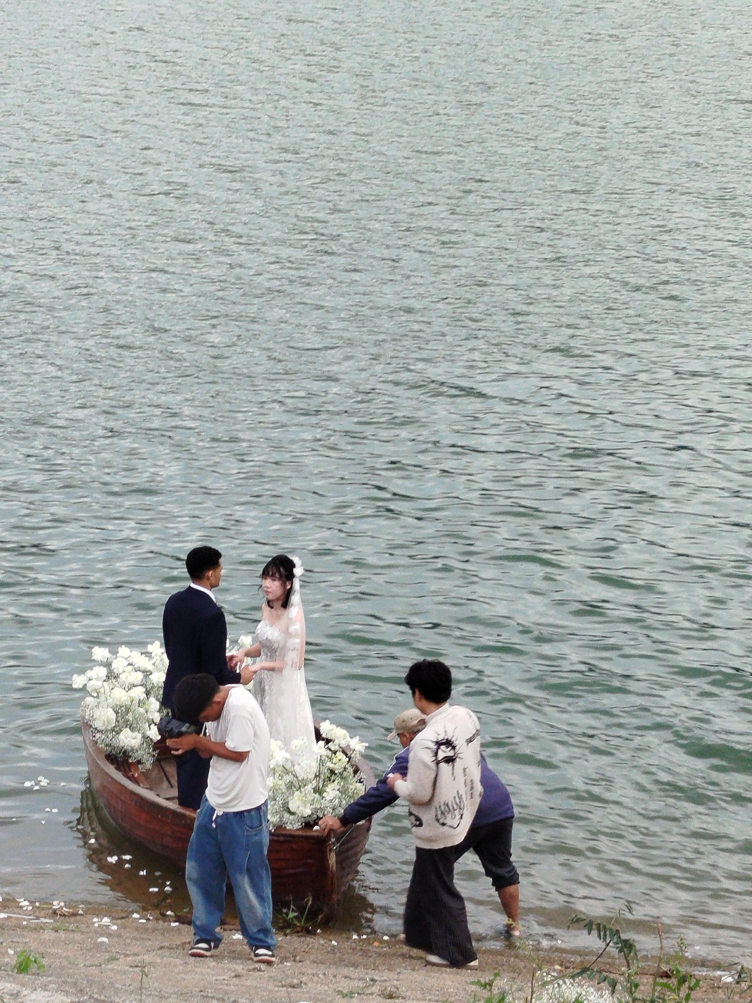 The image depicts a wedding photoshoot taking place on a boat in a body of water, likely a lake or reservoir, near Da Lat, Vietnam. A couple, dressed in wedding attire, stands in the boat which is adorned with white flowers. The groom is in a dark suit, and the bride wears a white wedding dress with a veil.
Several people are involved in the photoshoot. A photographer in a white t-shirt and blue jeans is crouched down, actively taking pictures of the couple with a camera. Another individual, possibly an assistant or second photographer, is standing near the front of the boat, and another person in a light-colored jacket and dark pants stands on the shore, interacting with someone near the boat. The boat itself is a traditional wooden rowboat, partially submerged in the shallow water at the edge of the shore.
The setting is a natural outdoor environment with a vast expanse of rippling water in the background. The shore is a mix of sand and gravel, with some sparse vegetation. The lighting suggests it might be daytime, possibly overcast or early morning/late afternoon, as there are no harsh shadows. The overall mood appears to be focused and professional, with the participants engaged in the task of capturing the wedding moments. There is no visible text in the image.
