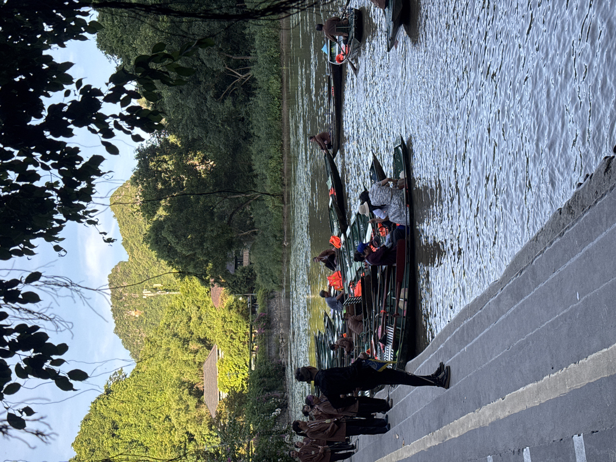 In Yên Sơn Ward, Vietnam, a group of tourists are lining up on a concrete embankment to board a fleet of traditional wooden boats, known as sampans, for a river tour. The boats are arranged in a line on the placid, rippling water, with passengers already seated in some. The tour guides, dressed in matching brown uniforms, are assisting the tourists. The background features lush green trees and karsts, characteristic of the Vietnamese landscape, with a few buildings nestled among the foliage. The sky is partly cloudy, and the sun casts a warm glow on the scene.