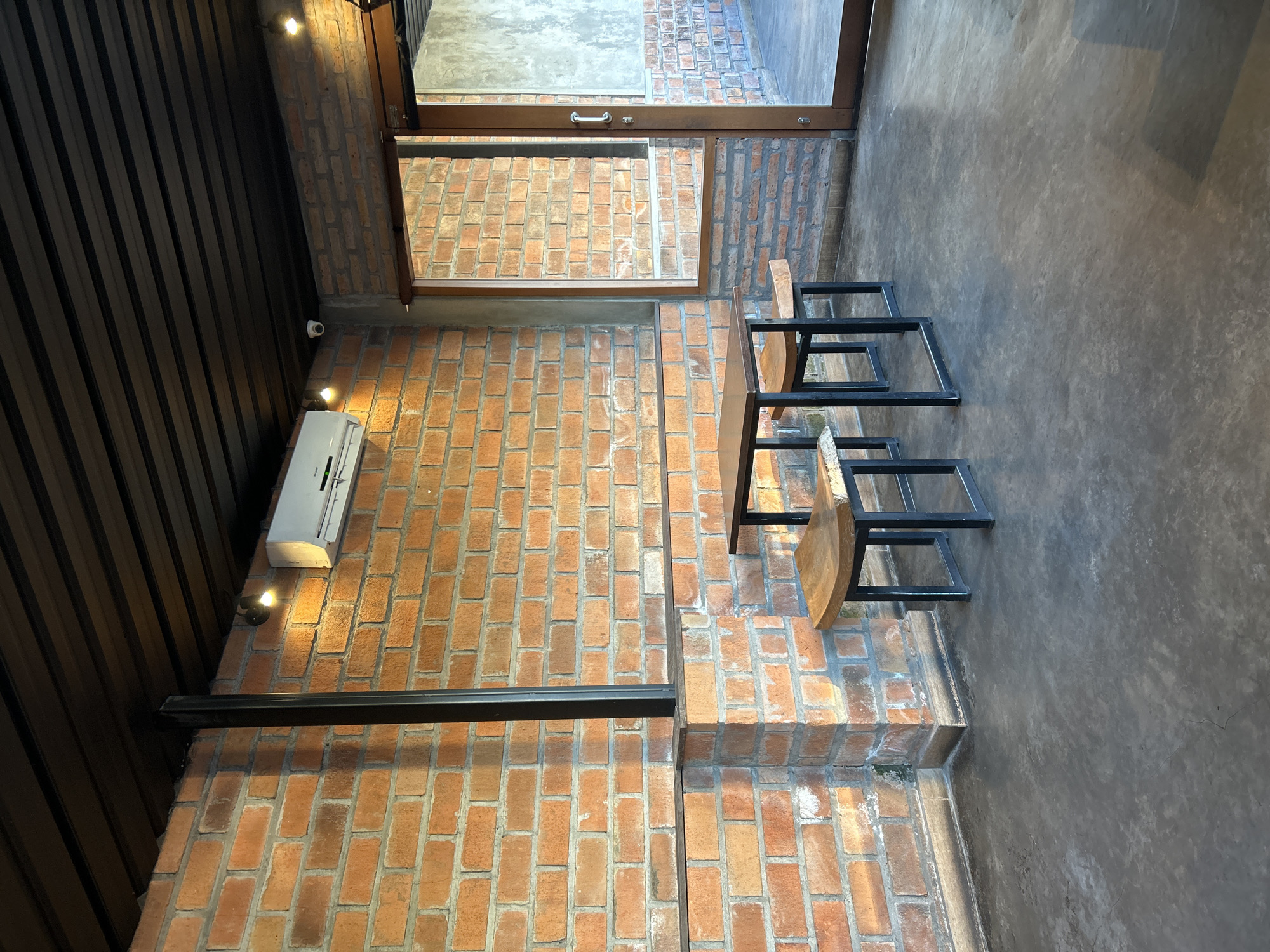 The image depicts an outdoor seating area of a cafe in Sukawati, Indonesia. A small, square table with a dark wooden top and black metal legs is set with two matching stools. The stools also feature dark metal legs and natural wood seats. The seating area is bordered by a brick wall on the left and a large glass-paned door and window on the right. Above the brick wall, a dark, slatted ceiling is visible. An air conditioning unit is mounted high on the brick wall, along with two small spotlights. A security camera is also visible near the ceiling. The floor is a smooth, gray concrete. The overall aesthetic is industrial-chic, with exposed brick and minimalist furniture.