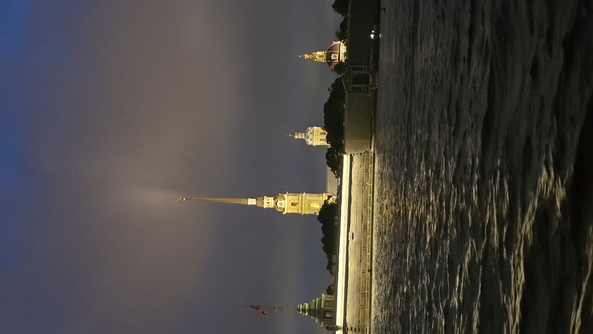 This nighttime, low-angle shot captures a view of the Peter and Paul Fortress in Saint Petersburg, Russia, from across the Neva River. The fortress, a prominent historical landmark, is illuminated by artificial lights, highlighting its distinctive architecture. The central feature is the Peter and Paul Cathedral's spire, a tall, slender structure topped with a golden angel. To the left of the spire, another tower with a pyramidal roof and a flag flying from its top is visible. To the right, a smaller, bulbous dome building also glows with light. The fortress walls, constructed from large, light-colored stones, form a solid barrier along the river's edge, with reflections of the lights shimmering on the dark, rippling water. The sky above is a moody gradient of deep blue and gray, suggesting overcast conditions or dusk. The overall scene conveys a sense of historical grandeur and quiet nighttime ambiance.