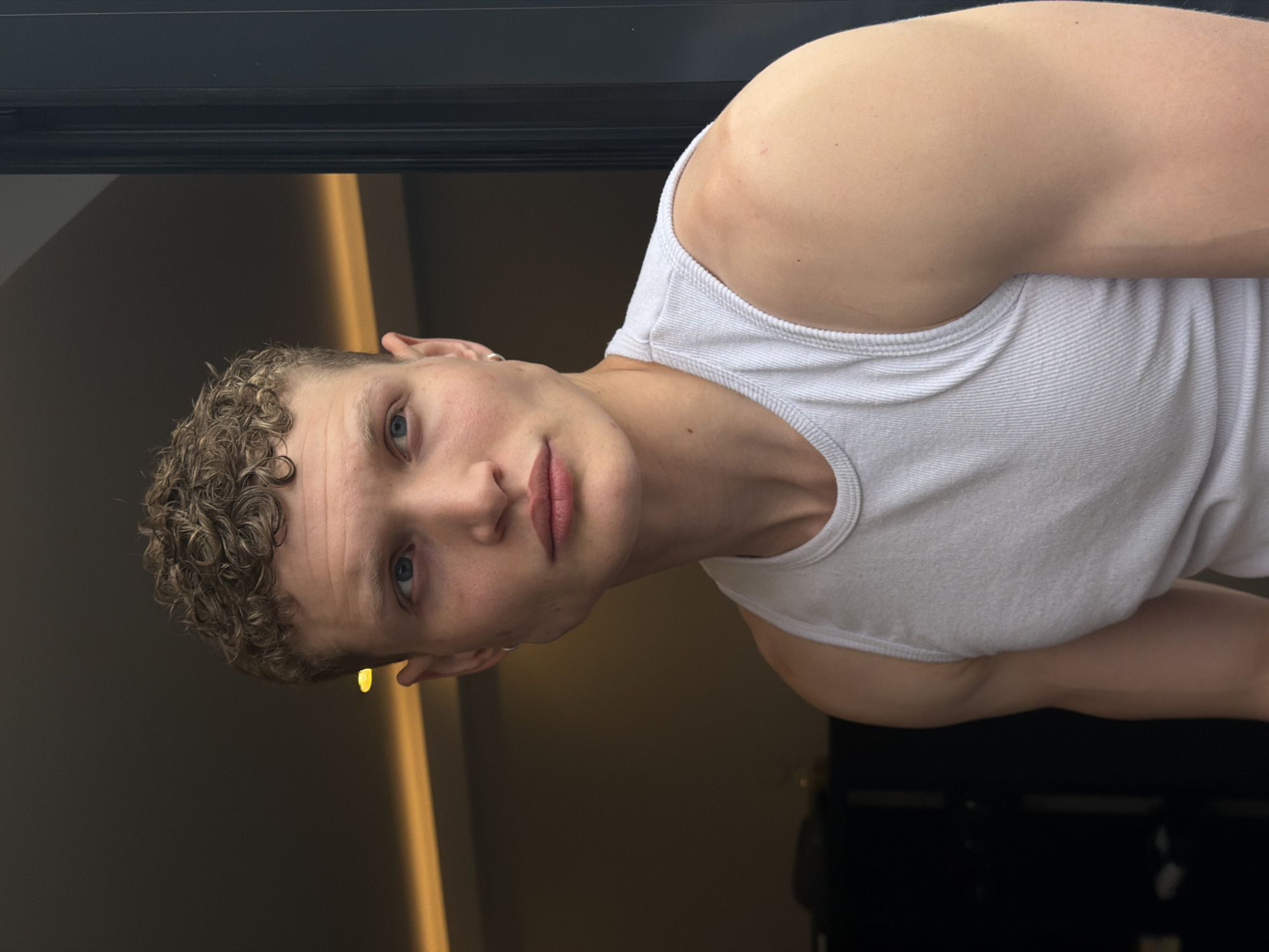 A young Caucasian male with curly blonde hair is wearing a white ribbed tank top. He is positioned indoors, likely near a window or doorway, as evidenced by the dark frame at the top left. A vertical light fixture is visible in the background to the left. The scene has a dark interior, with glimpses of possibly dark furniture in the lower right corner. The location is not discernible from the image. The male is looking directly at the camera with a slightly tilted head.