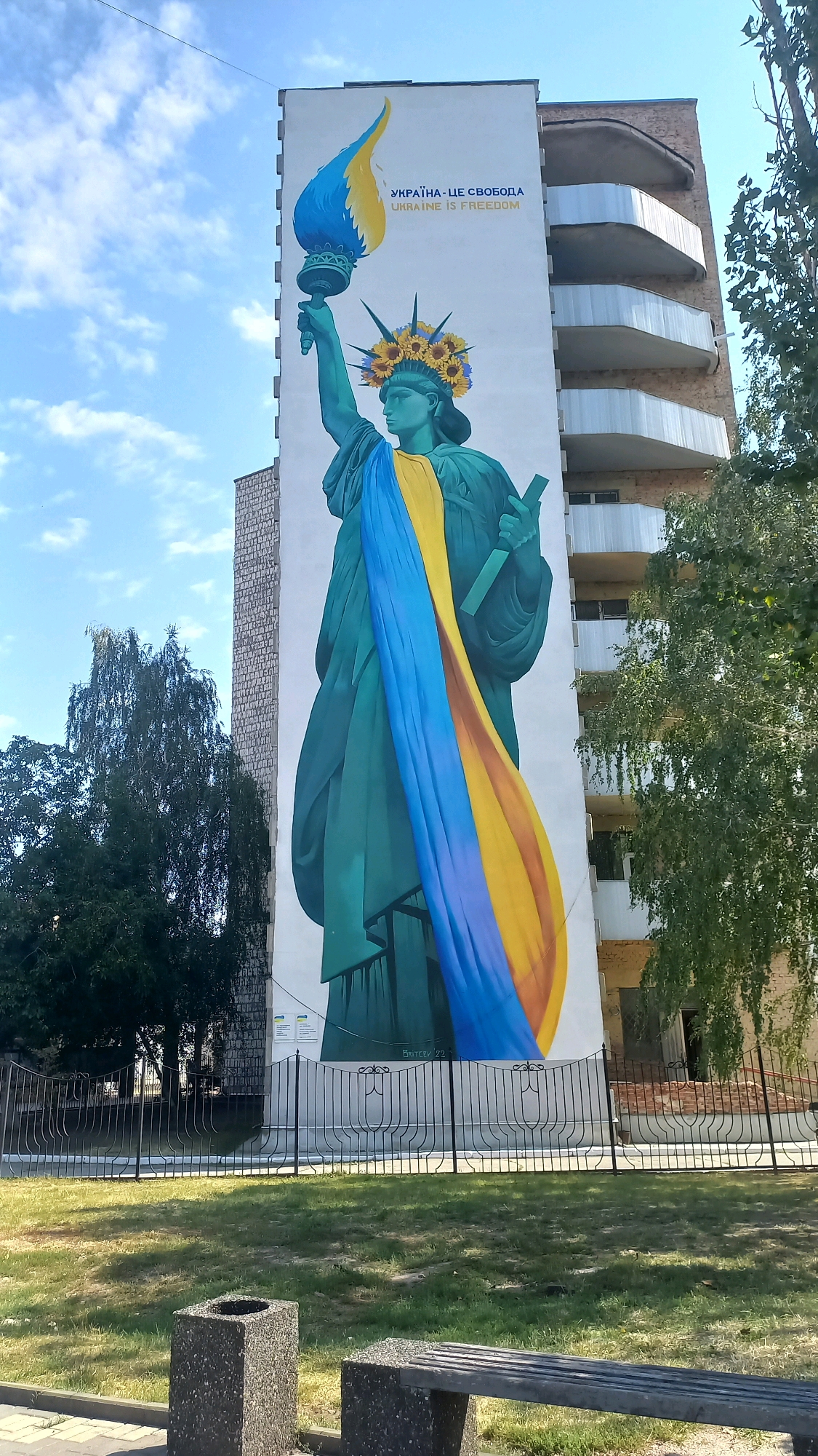 This is a photograph of a mural on the side of a building in Kropyvnytskyi, Ukraine. The mural depicts a stylized version of the Statue of Liberty holding a torch aloft. The torch flame is rendered in the colors of the Ukrainian flag, blue and yellow. The statue's headdress is adorned with sunflowers, and a draped sash across its body also features the blue and yellow of the Ukrainian flag. Above the mural, text reads "УКРАЇНА - ЦЕ СВОБОДА" and "UKRAINE IS FREEDOM." The building itself is multi-story with balconies. In the foreground, a black metal fence separates the mural from a grassy area with a bench and a concrete trash receptacle. Trees are visible to the left and right of the building. The sky is blue with scattered white clouds. The mural appears to be located on a building on a street, with other buildings visible in the background. The overall scene conveys a message of Ukrainian freedom and resilience.