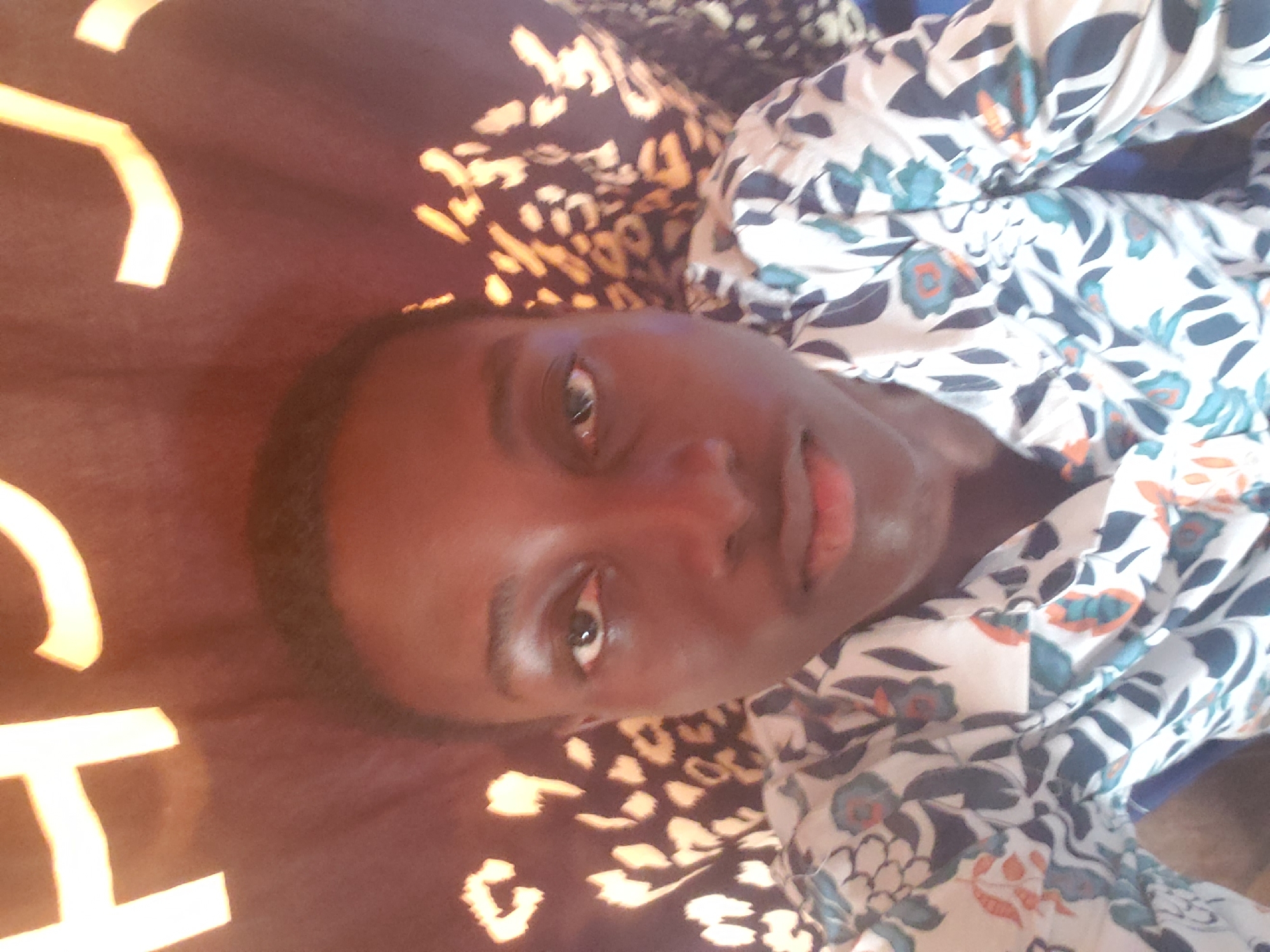 A person is lying on a surface. The person has dark skin and is wearing a white shirt with a floral pattern. The surface appears to be a bed with a patterned bedsheet. No information is available to identify the location as Alalubosa, Nigeria or any specific environment.