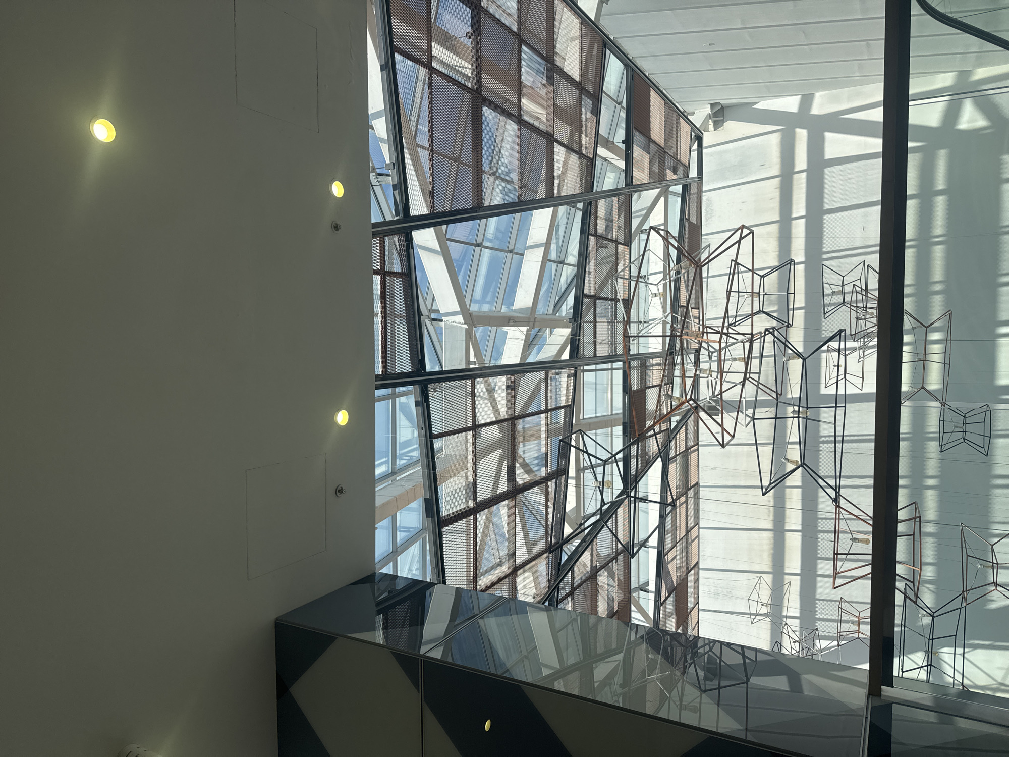 This is a photograph of the interior of the Grand Mall in Plovdiv, Bulgaria. The image captures the atrium space, looking upwards towards the ceiling and a large skylight. The ceiling is white and features several recessed light fixtures and what appears to be a smoke detector. Below the skylight, a complex geometric sculpture made of wire frames hangs suspended from the ceiling. The skylight itself is constructed with a grid of glass panels and a metal mesh overlay, allowing natural light to filter through and cast shadows on the interior walls. The walls on the right side of the image are a light, textured white, with vertical paneling. On the left, a large, angular column clad in reflective dark and light grey panels is visible, reflecting the interior architecture. The overall impression is one of modern, spacious interior design.