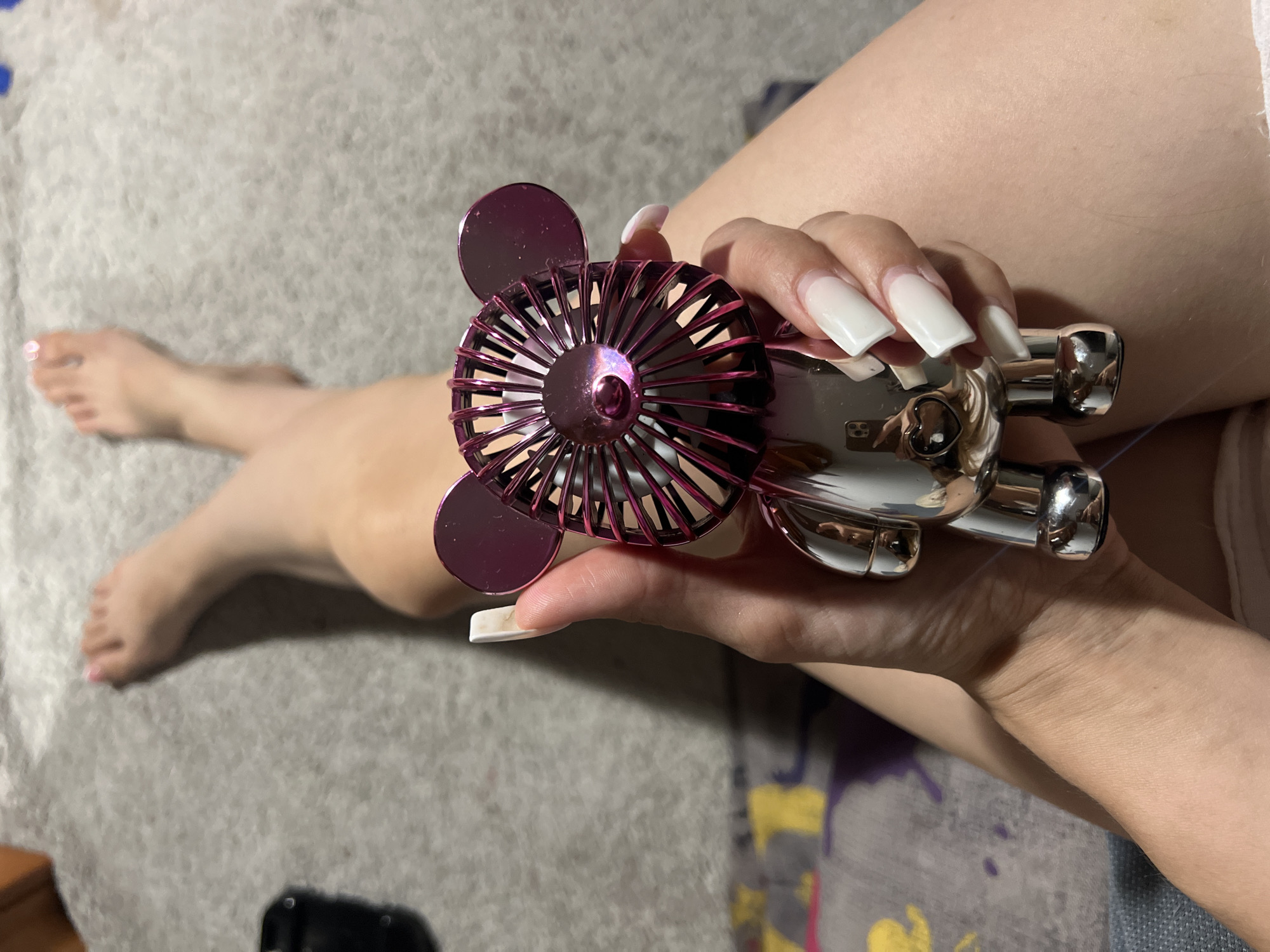 A close-up, high-angle shot shows a person's legs and a hand holding a small, metallic fan shaped like a cartoon bear. The fan is pink and silver, with a fan blade in the head section and round ears. The person's feet are visible in the background, with painted toenails. The hand holding the fan has long, square, white acrylic nails. The scene appears to be indoors, with a gray carpeted floor and a colorful blanket or fabric visible beneath the legs. The lighting suggests an artificial light source. The image is focused on the hand and fan, with the legs and feet slightly out of focus.