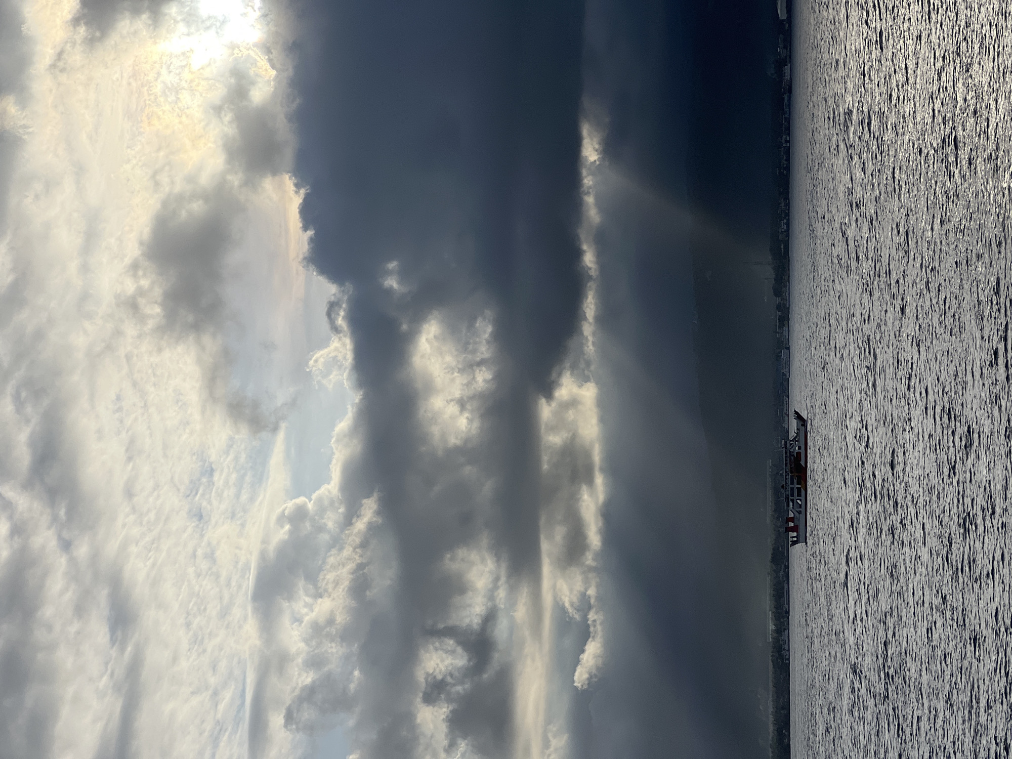 The image depicts a body of water, likely a sea, with a ship sailing along the coastline. On the coastline, there are buildings and greenery, possibly indicative of a city. In the sky, there are dramatic cloud formations with sunlight breaking through, creating a contrast between light and shadow. The image appears to be taken during the day.
