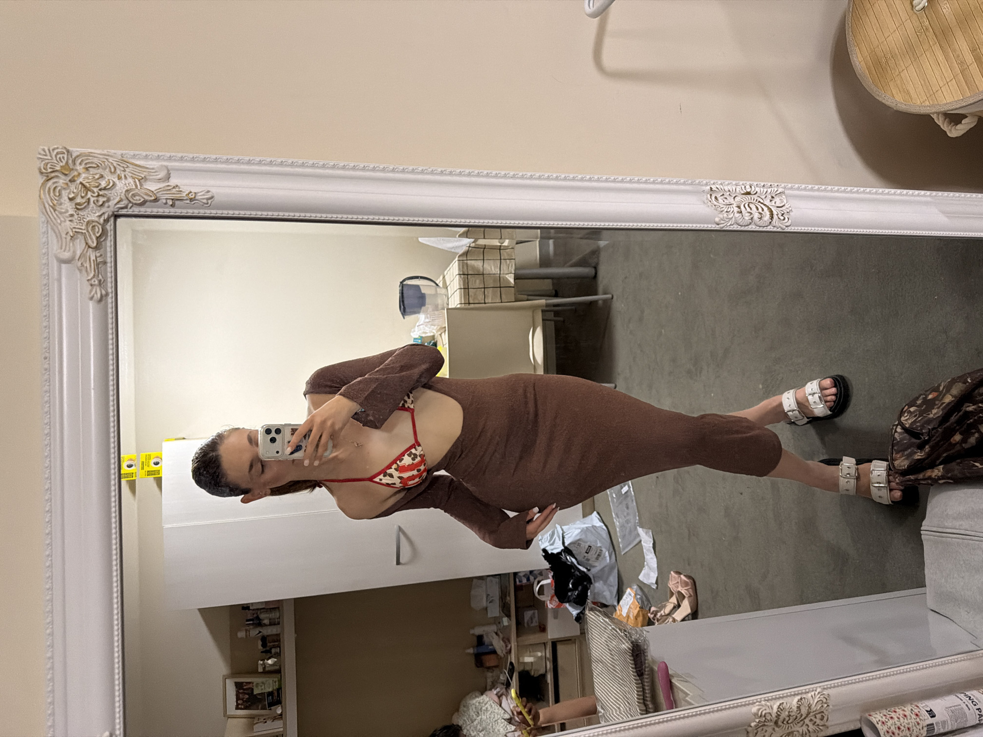 A woman is reflected in a large, ornate white-framed mirror. She is wearing a long-sleeved brown dress with a cutout at the chest, revealing a red-and-white patterned bikini top. She is holding a smartphone and appears to be taking a selfie. The reflection reveals a cluttered room with various objects, including furniture, bags, and miscellaneous items on shelves and on the floor. There is no visible indication of the location being Sofia, Bulgaria.