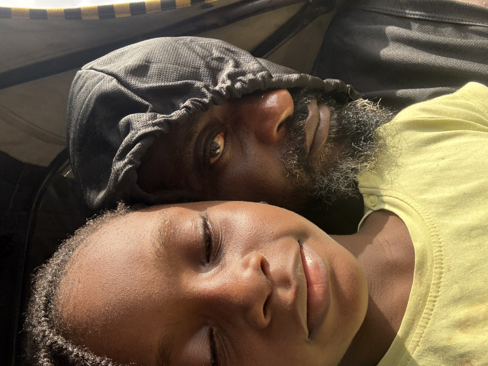 A close-up shot depicts an adult male wearing a dark grey hooded sweatshirt with the hood up, lying down with his head resting above a young girl who is wearing a light yellow t-shirt. The man has a dark complexion and a grey-streaked beard. His eyes are open and he appears to be gazing at the camera. The young girl has her eyes closed. The background is blurry and indistinct, with what appears to be a canopy above. The scene is illuminated by bright, natural light, casting shadows and highlighting textures on the skin. It is not possible to confirm that the location is Ado, Nigeria, from this image.