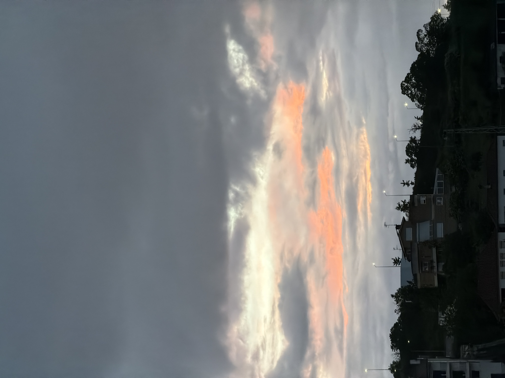 The image shows a sky at dusk with orange and white clouds. The view includes a hillside with houses, trees, and streetlights. This is in Bragança, Portugal.