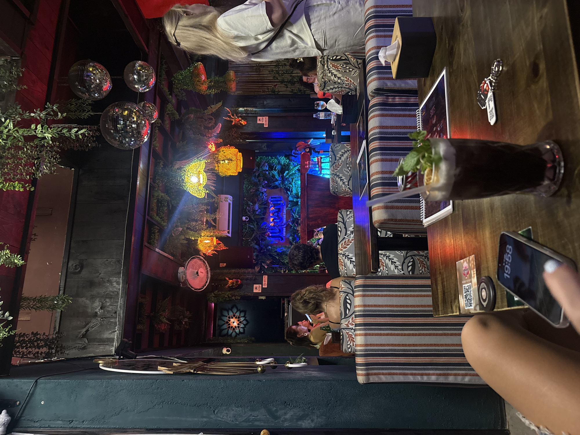 The image shows the interior of a bar or restaurant in Nha Trang Ward, Vietnam. A dimly lit atmosphere is enhanced by decorative disco balls, hanging foliage, and various decorative lighting fixtures. Several people are seated at tables with striped cushions, some appearing to be engaged with each other, while others are occupied with their phones. A cocktail is placed on a table next to a menu, and various objects like tissues and keychains are also on the table. The background features lush greenery and colored lighting, contributing to a vibrant and somewhat enclosed setting.