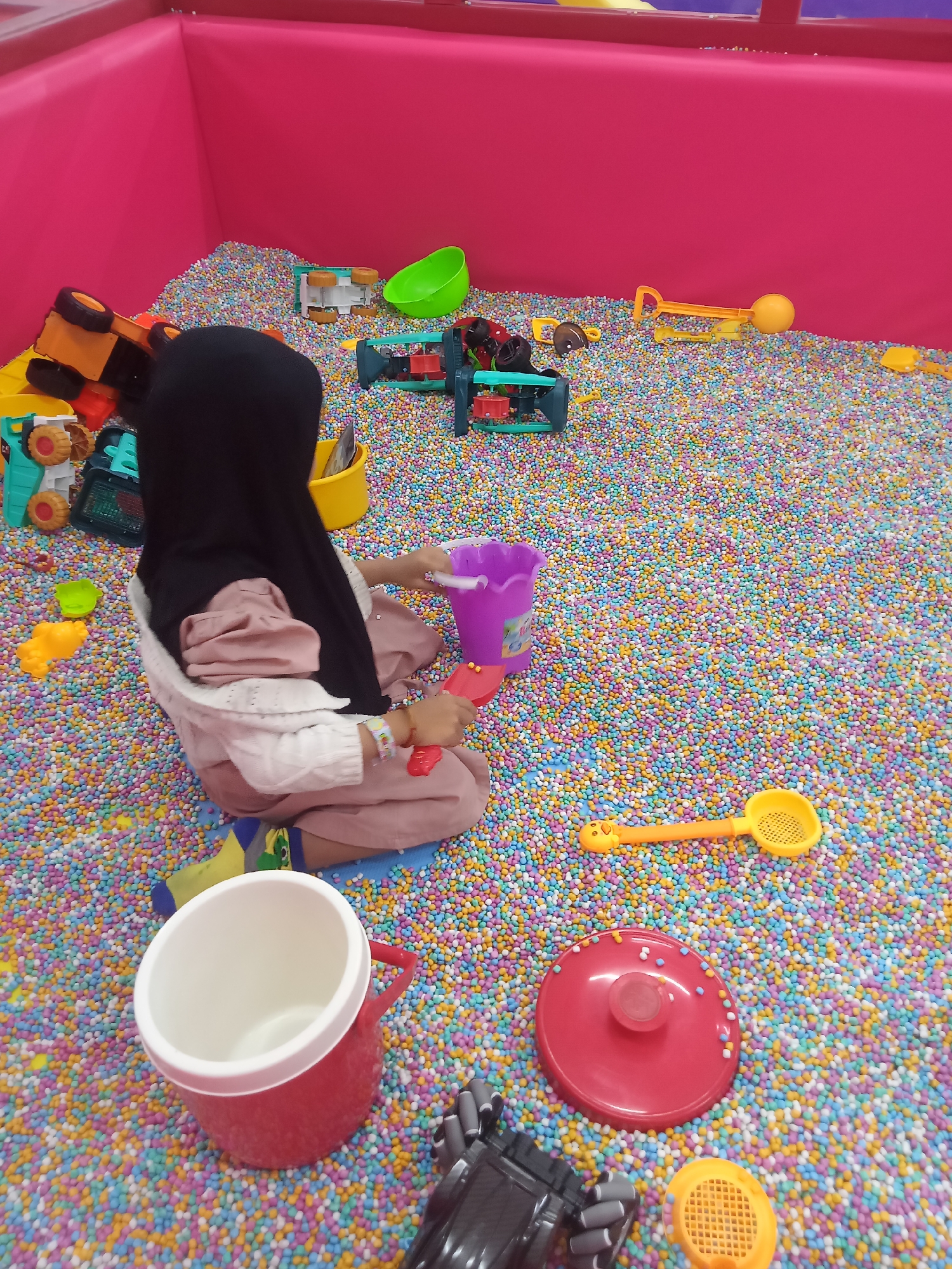 A young child, wearing a black hijab and pink clothing, sits in a ball pit filled with colorful small balls. The child is facing away from the camera and appears to be playing with a purple bucket and a red scooper-like toy. Scattered throughout the ball pit are various plastic toys, including trucks, a green bucket, and yellow scoops. The ball pit is enclosed by bright pink walls. The environment suggests an indoor play area, possibly a children's entertainment center. The image is taken from a slightly elevated angle. The location is in Boyolali, Indonesia.