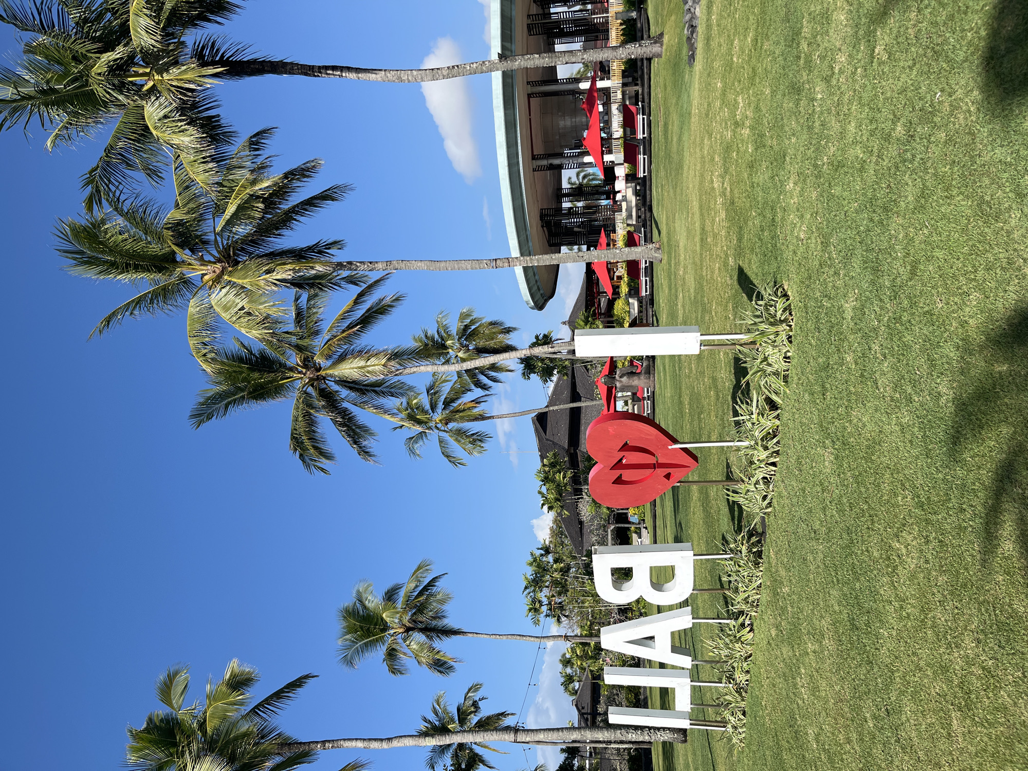 A bright, sunny day in Kuta Selatan, Indonesia, with a clear blue sky and scattered white clouds. In the foreground, a lawn displays large white letters spelling "BALI" and a red heart shape in between the "A" and "L." Behind the sign, several palm trees tower over the scene. In the middle ground, a resort area is visible, featuring lounge chairs and red umbrellas, with a modern, open-air building in the background. The overall atmosphere is tropical and inviting.