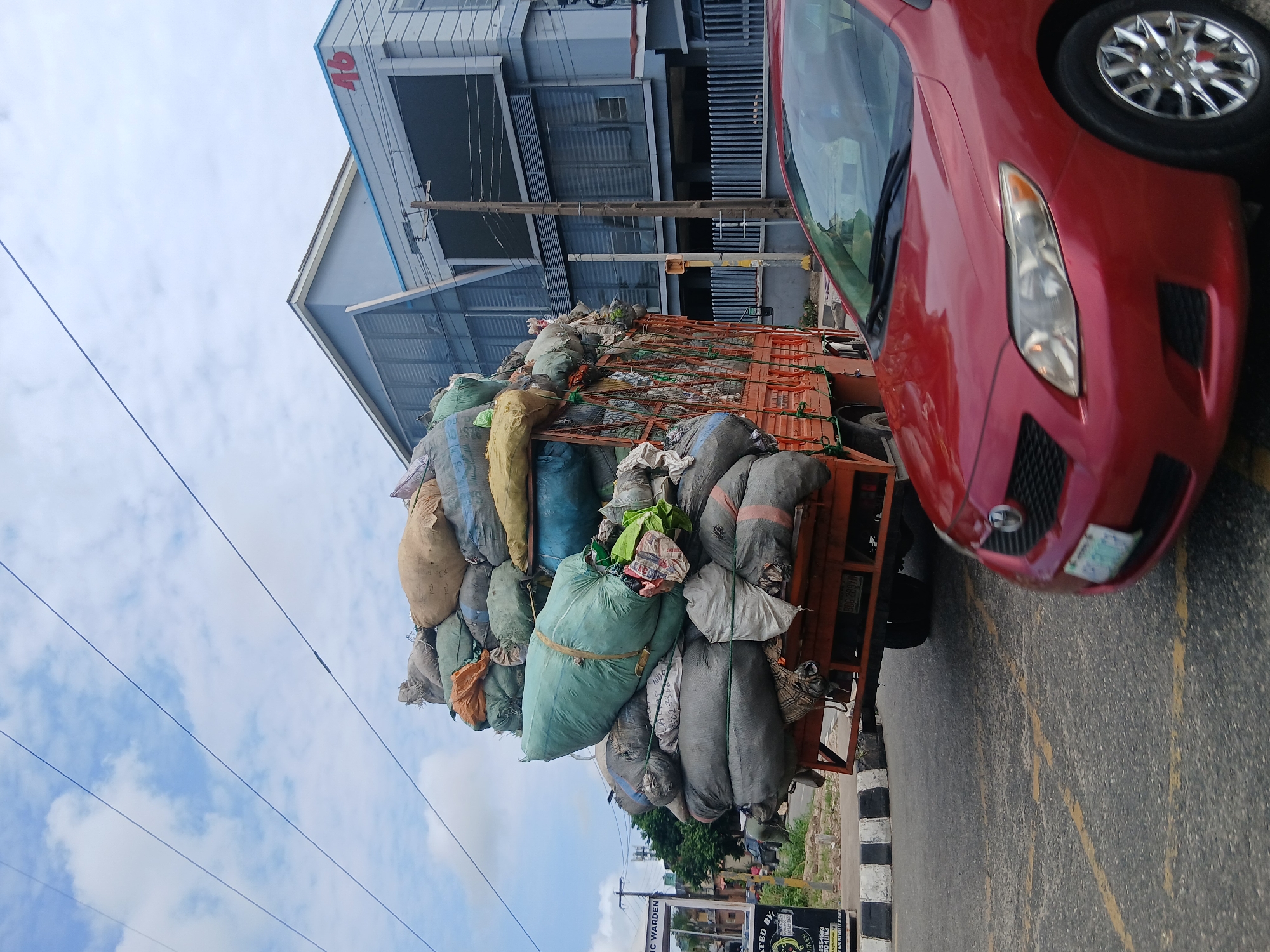 In Animashawun, Nigeria, a large truck is heavily loaded with numerous large sacks, appearing to be filled with various materials. The truck is red with an orange flatbed, and the sacks are tied down with ropes. Some sacks are green, while others are beige, gray, and blue. The truck is positioned on a road, with a red car in the foreground also on the road. The car is a sedan with a license plate that is partially visible. In the background, there is a modern building with a sign that reads "46". The sky is cloudy. There are utility poles and wires visible, as well as some trees and a street sign.